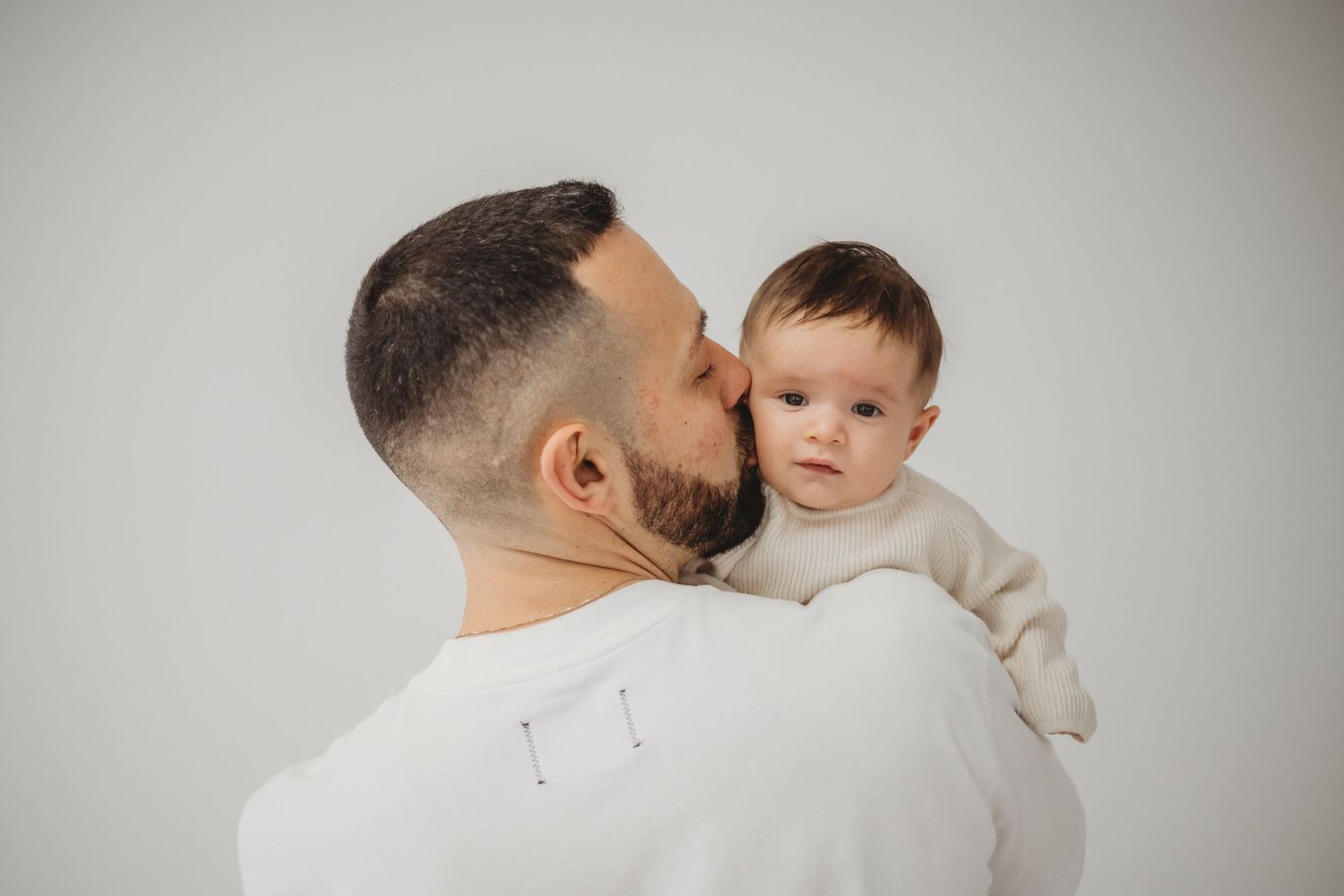 Bearded father kissing baby on cheek while infant looks at camera.