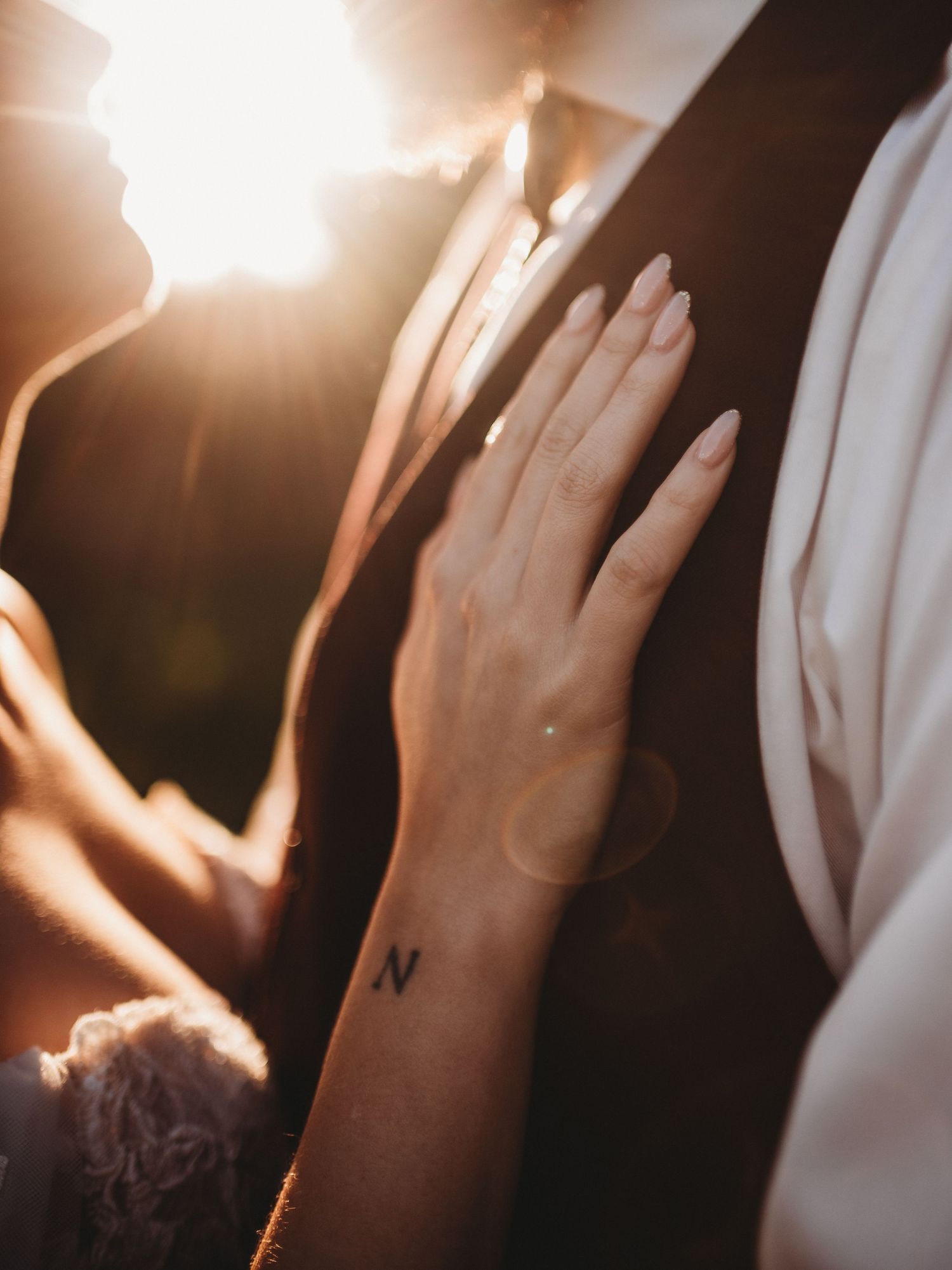 Close-up of bride's hand with letter N wrist tattoo resting on groom's vest in warm golden sunlight.
