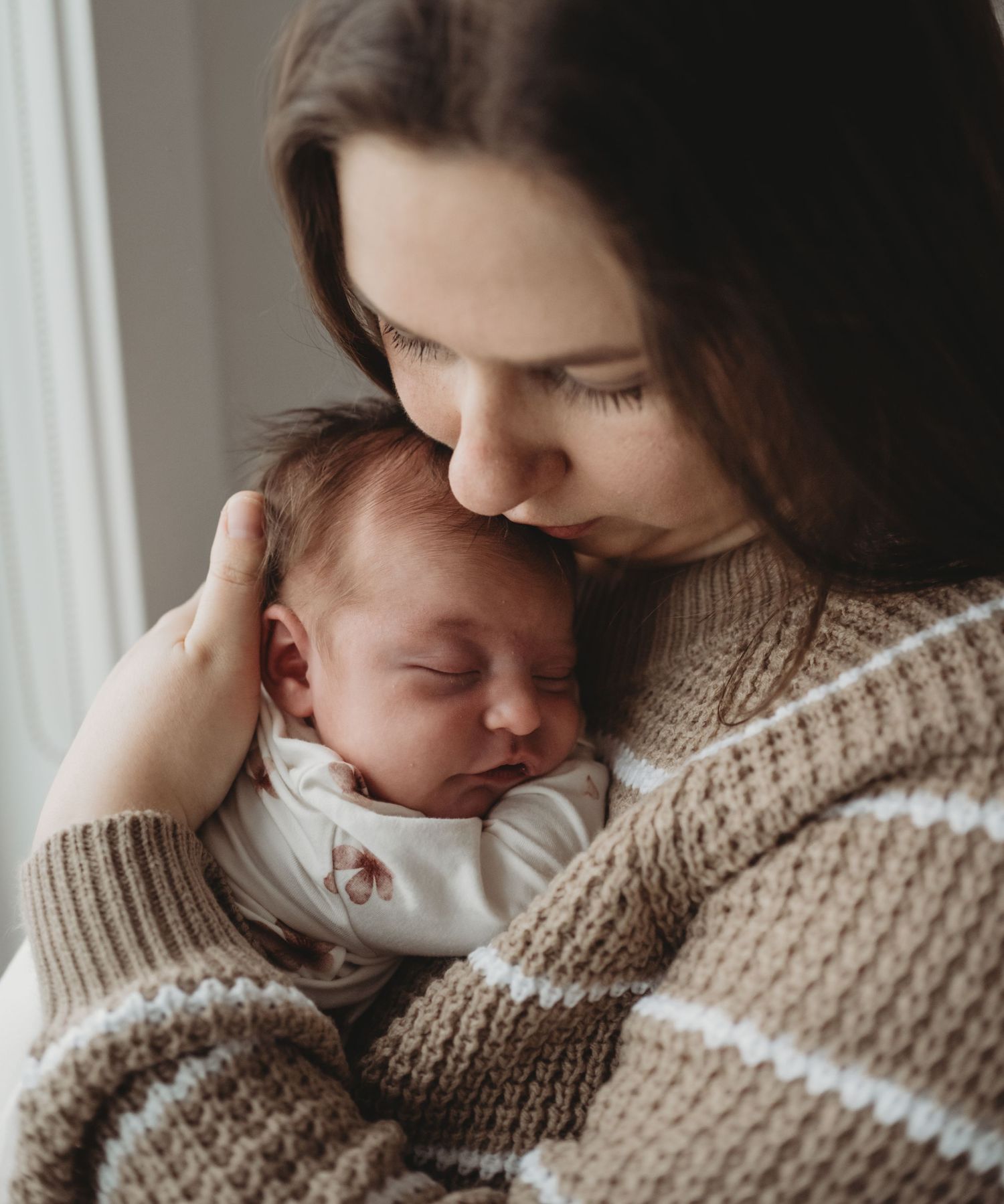 A brunette mother in a striped sweater gently kisses her sleeping newborn baby.