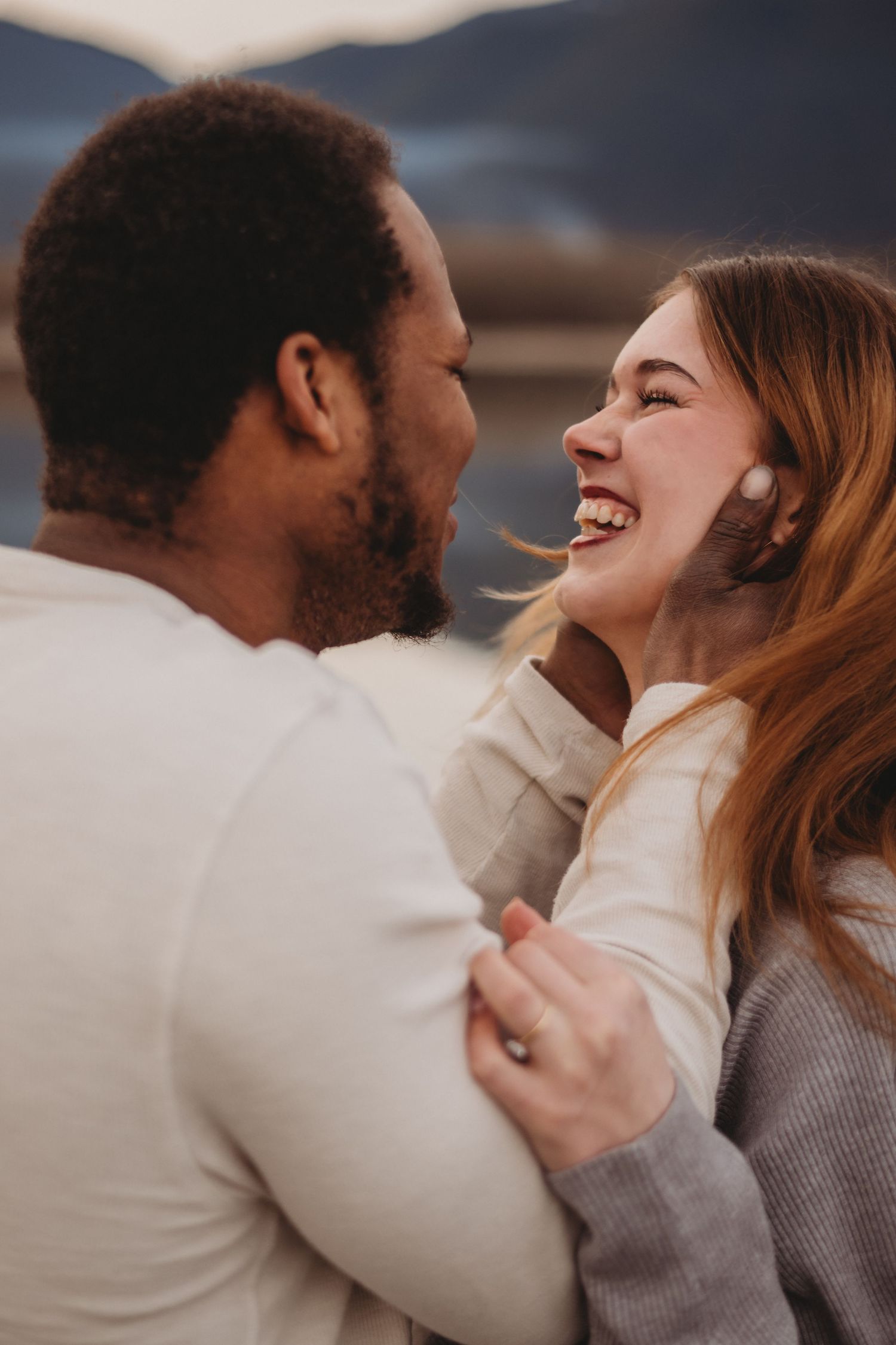 Happy interracial couple laughing together near a mountain lake during engagement photos.