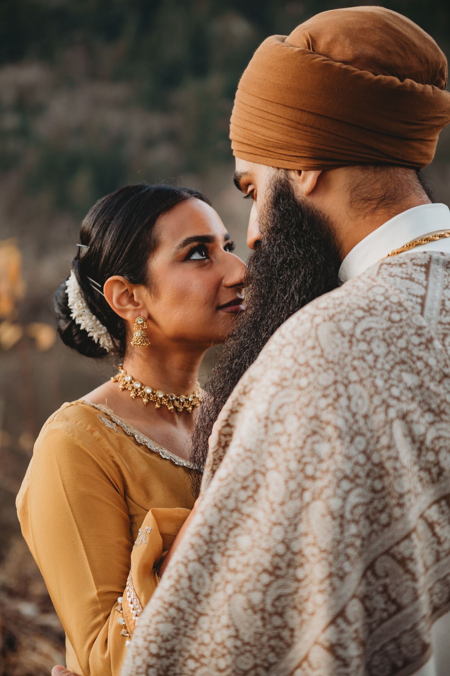Sikh bride adorned with gold necklace and jhumkas gazes lovingly at bearded groom in amber dastar.