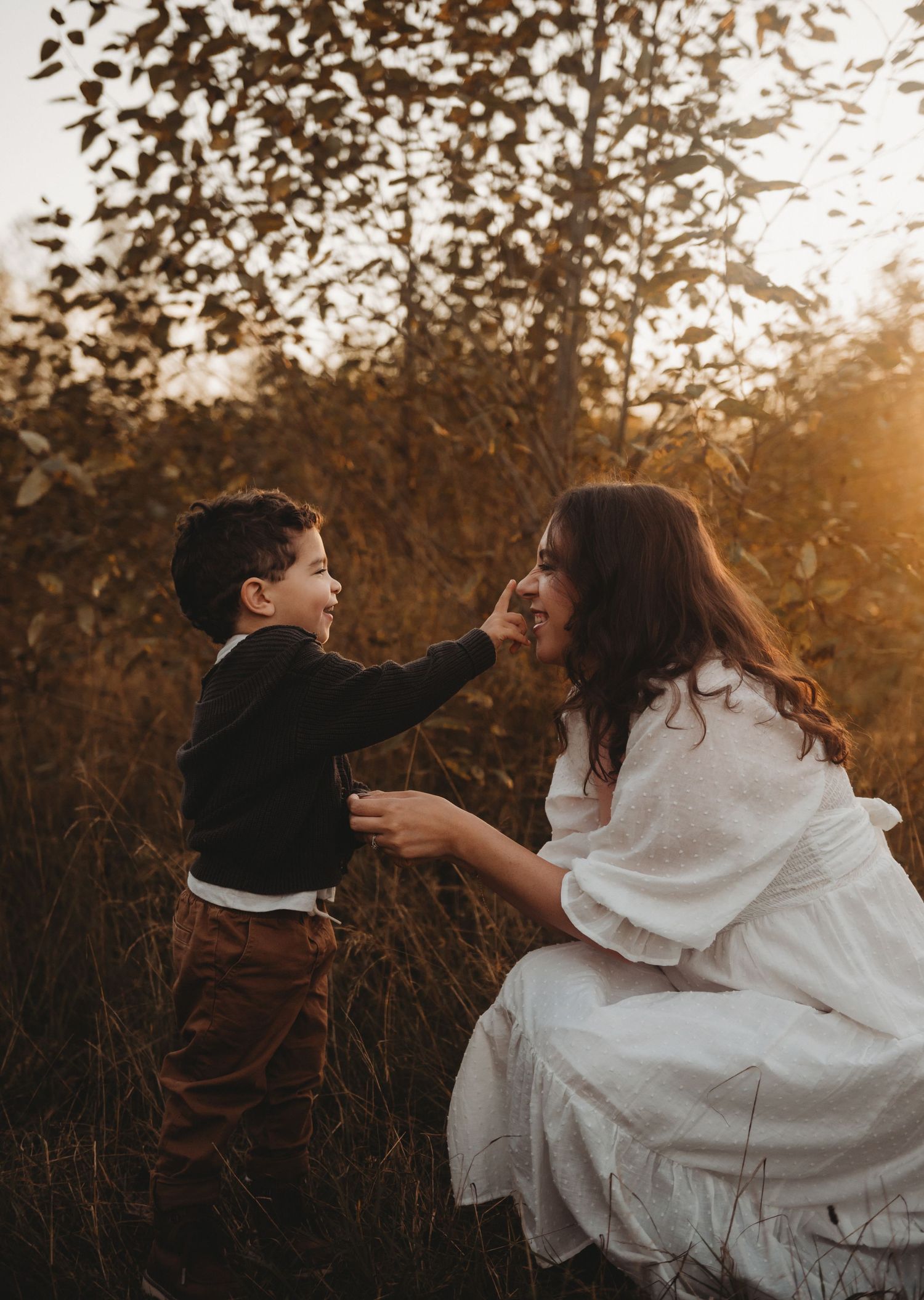 Young boy playfully touching his smiling mothers nose while she crouches in a sunlit autumn field.