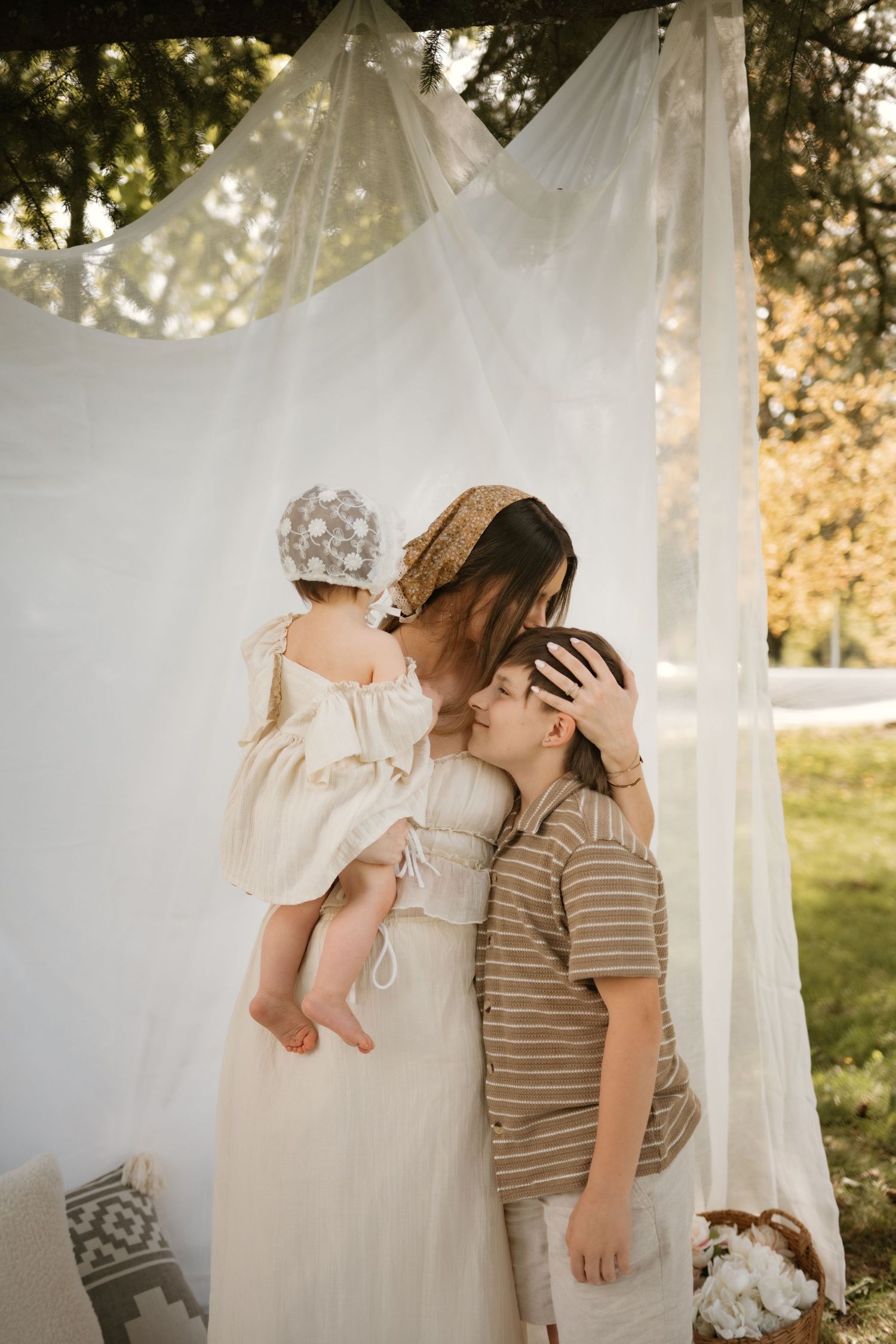 Mother holding baby and embracing young son under white draped canopy in outdoor setting.