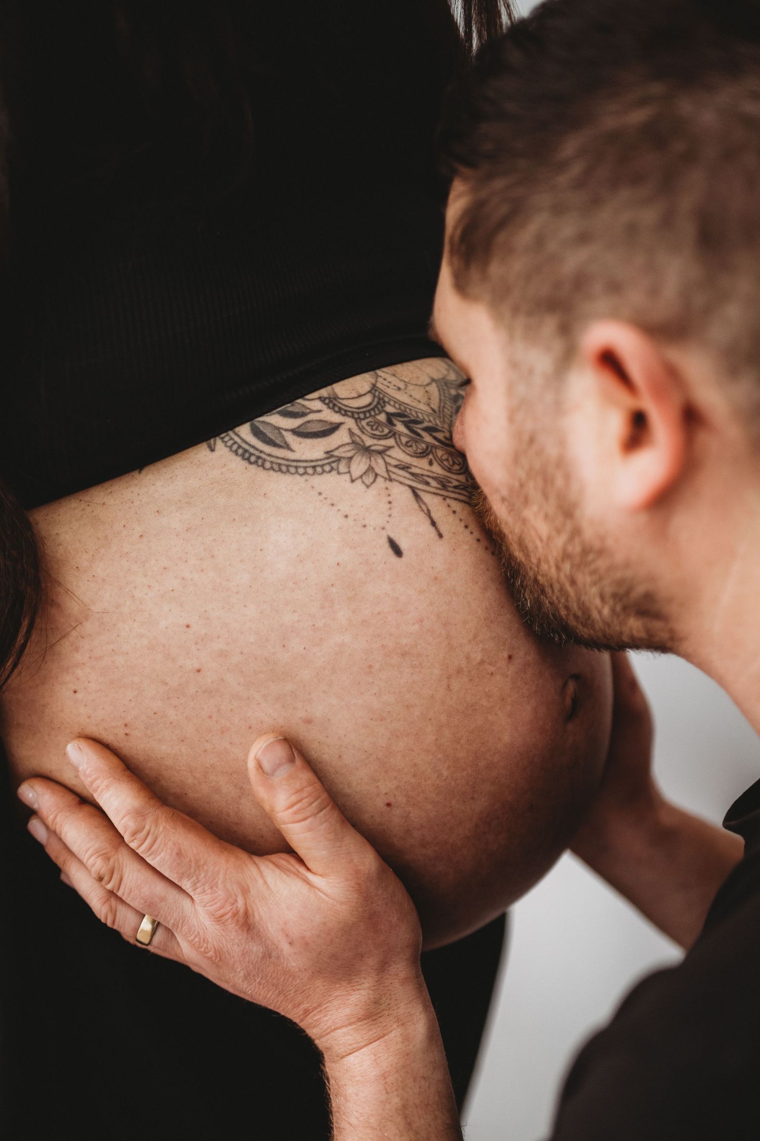 Close-up of man kissing pregnant belly with intricate mandala tattoo.