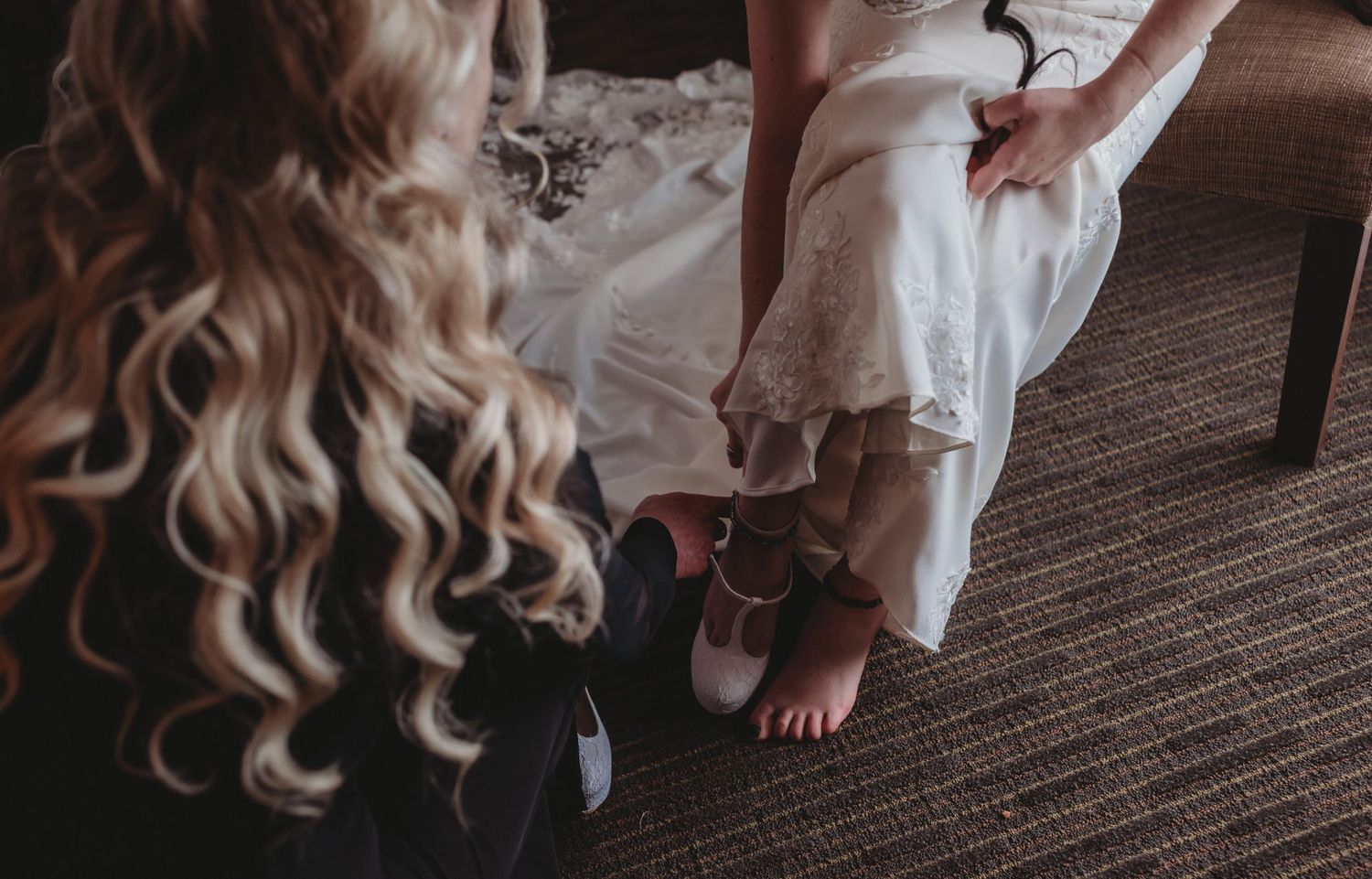 Bridesmaid with curly blonde hair helping bride with her shoes during wedding day preparations.