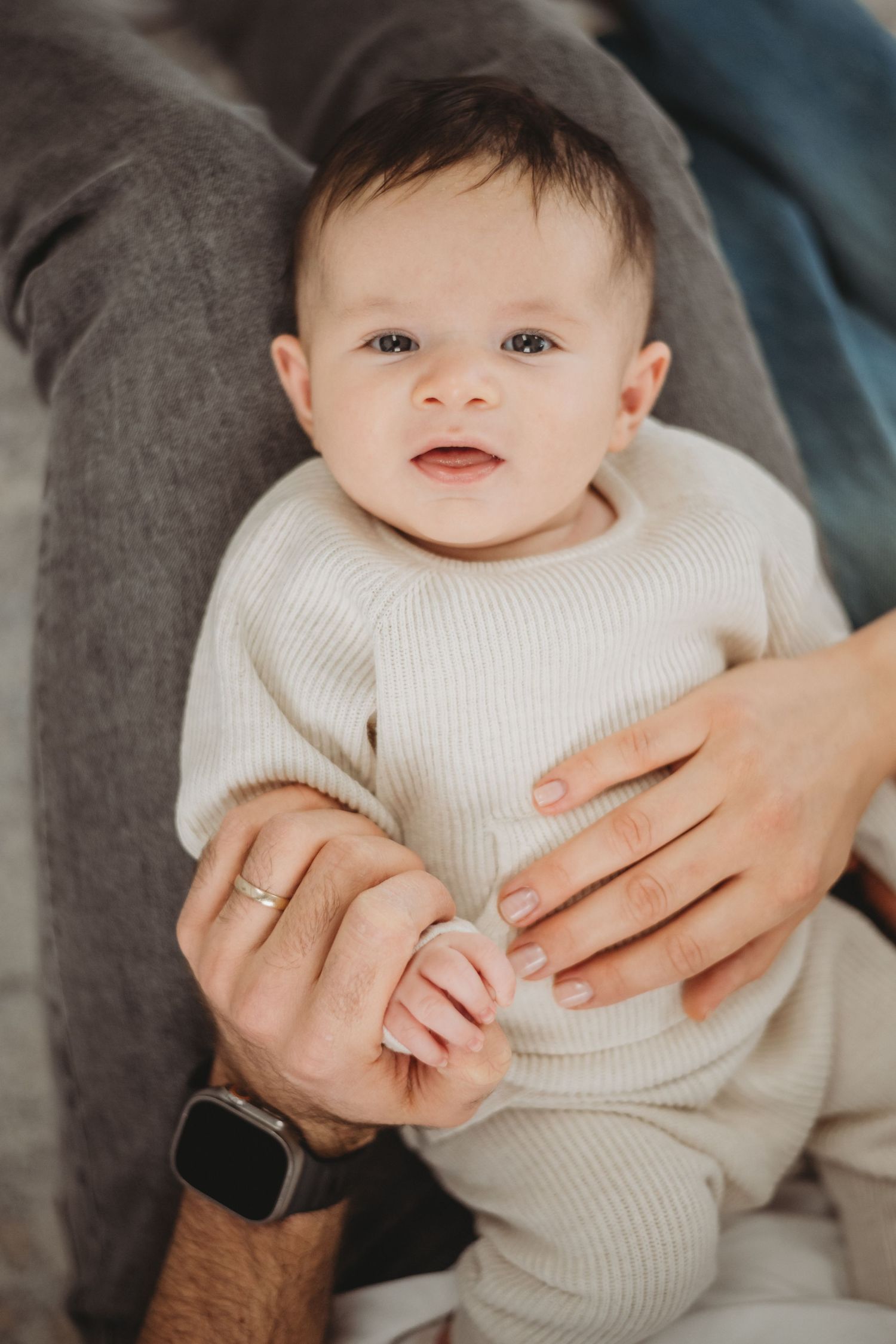 Alert dark-haired baby in cream knit outfit held by parents, looking at camera.
