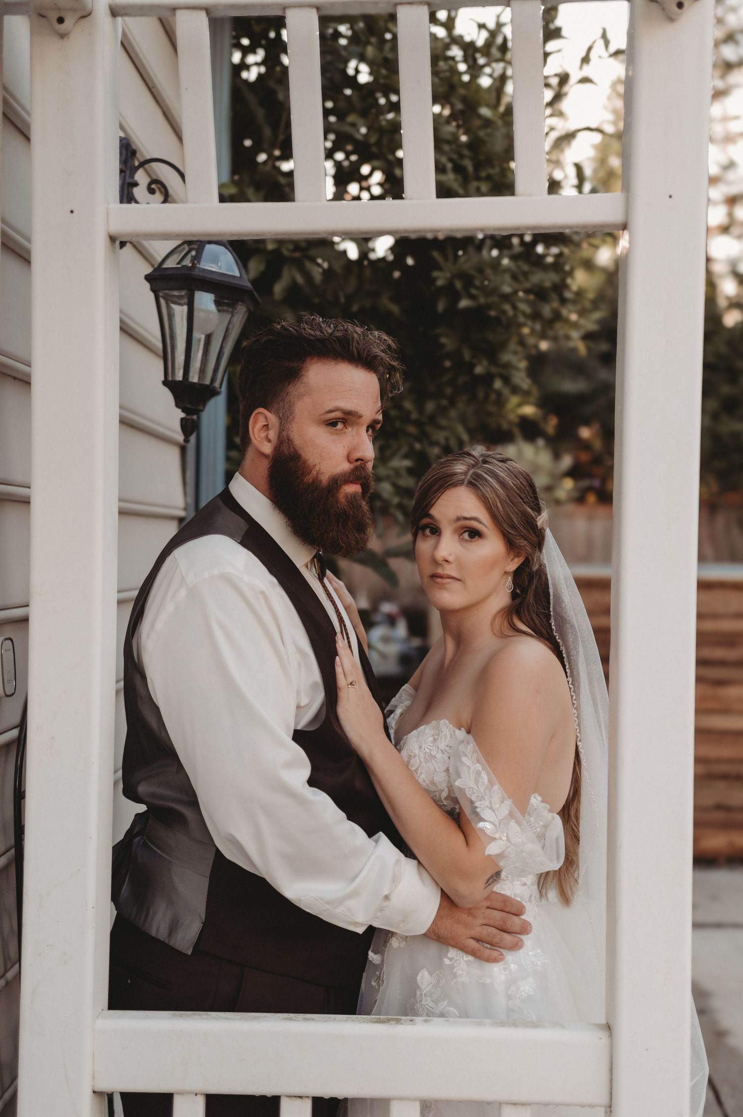 A bearded groom in a vest and bride in a lace gown pose elegantly in a white garden gazebo.