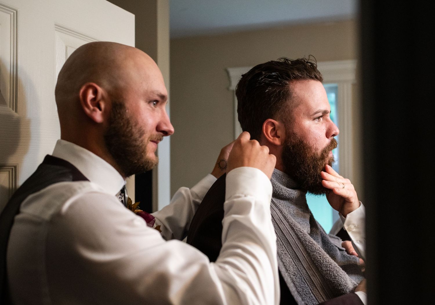 Bald groomsman helps bearded groom adjust his teal tie while getting ready indoors.