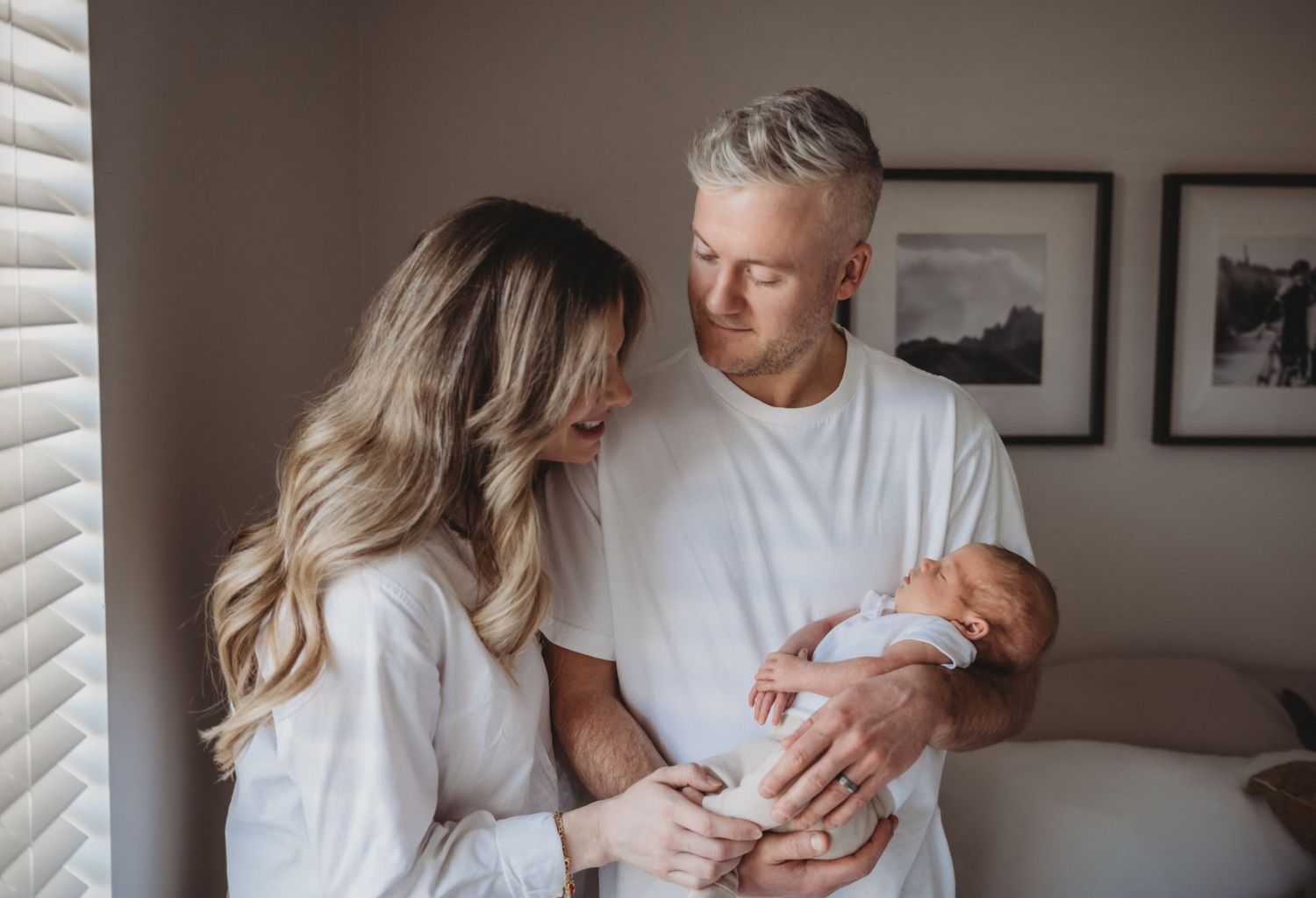 Young couple in white outfits lovingly gazing at newborn baby held in fathers arms indoors.