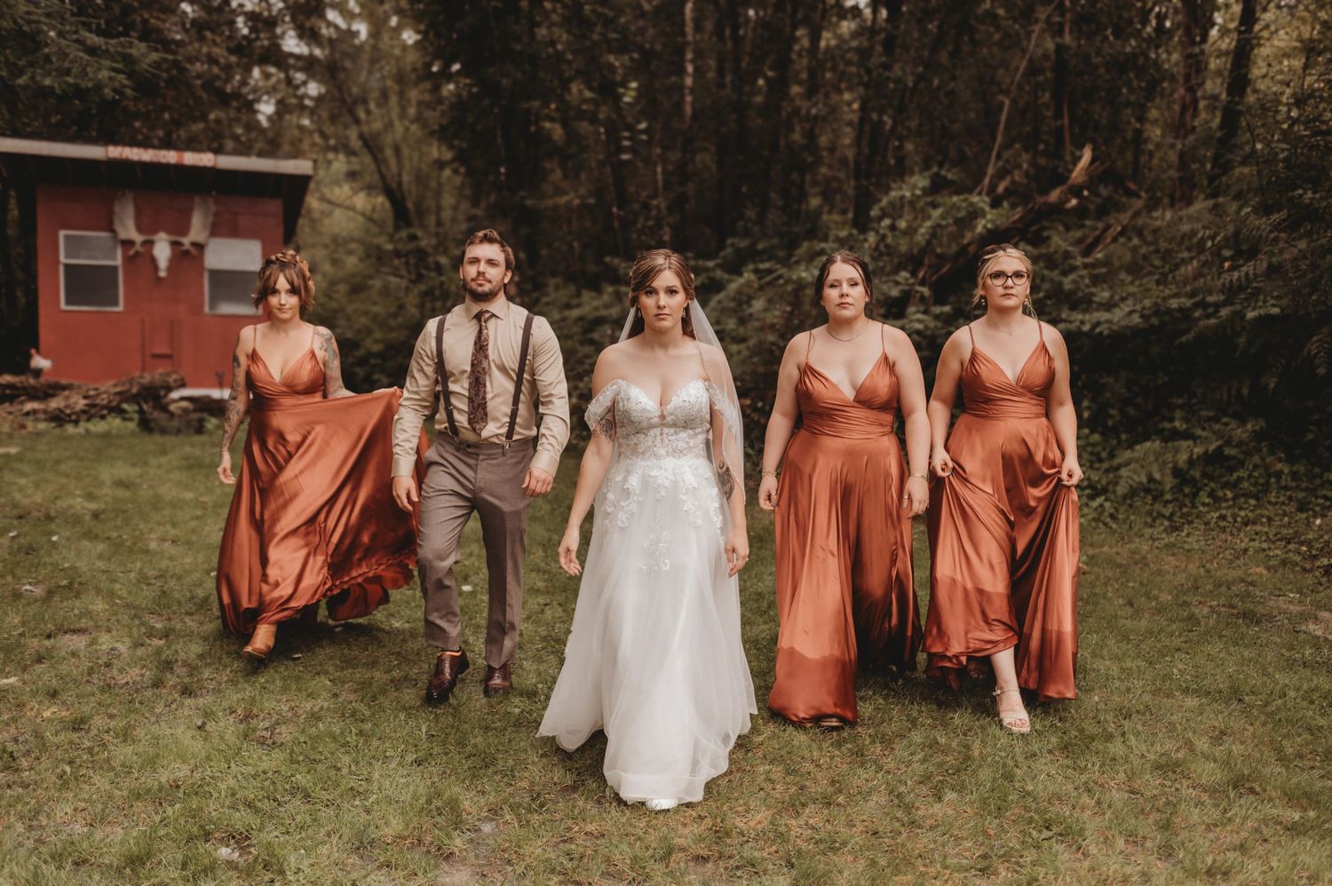 Bride in white lace gown walks confidently with bridesmaids in flowing rust orange dresses outdoors.
