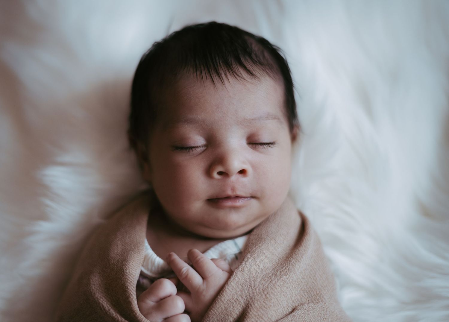 Peaceful sleeping newborn baby wrapped in beige swaddle on white fluffy blanket.
