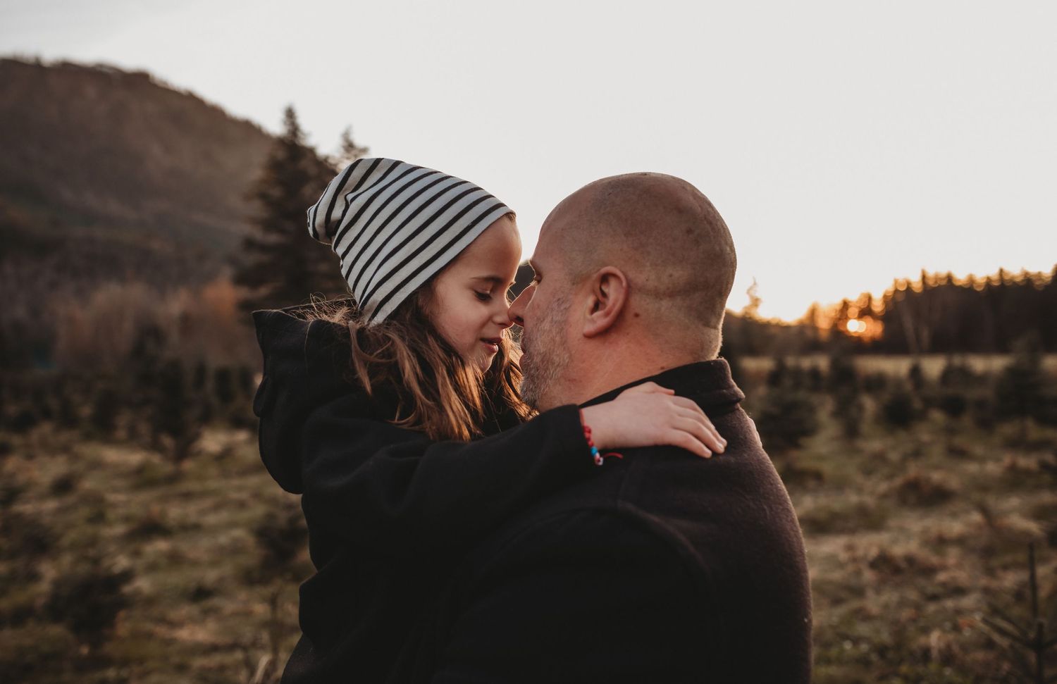 A bald father tenderly embraces his young daughter in a striped beanie at golden hour outdoors.