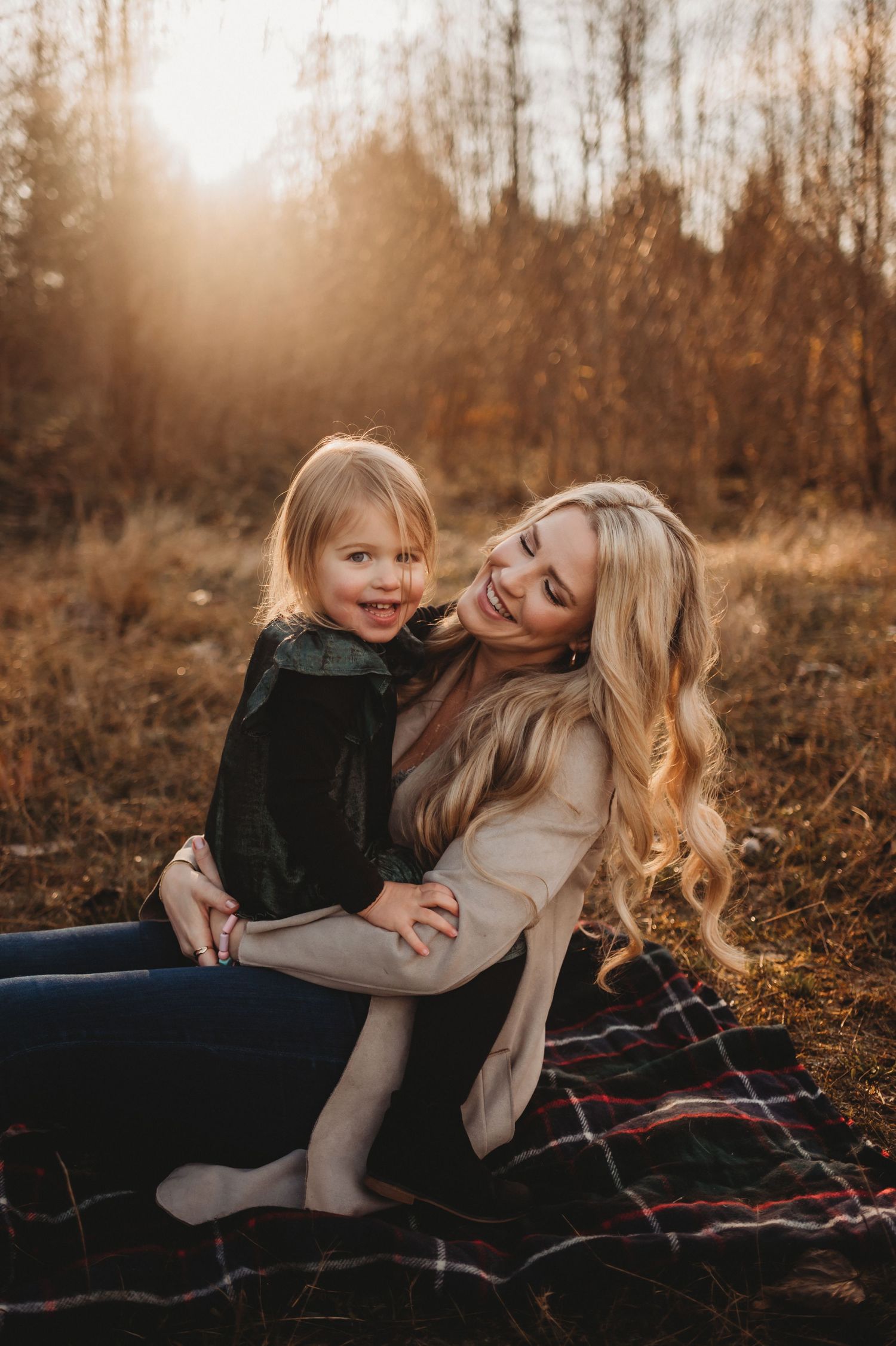 A blonde woman smiles warmly while holding a laughing toddler girl outdoors during golden hour in an autumn field.