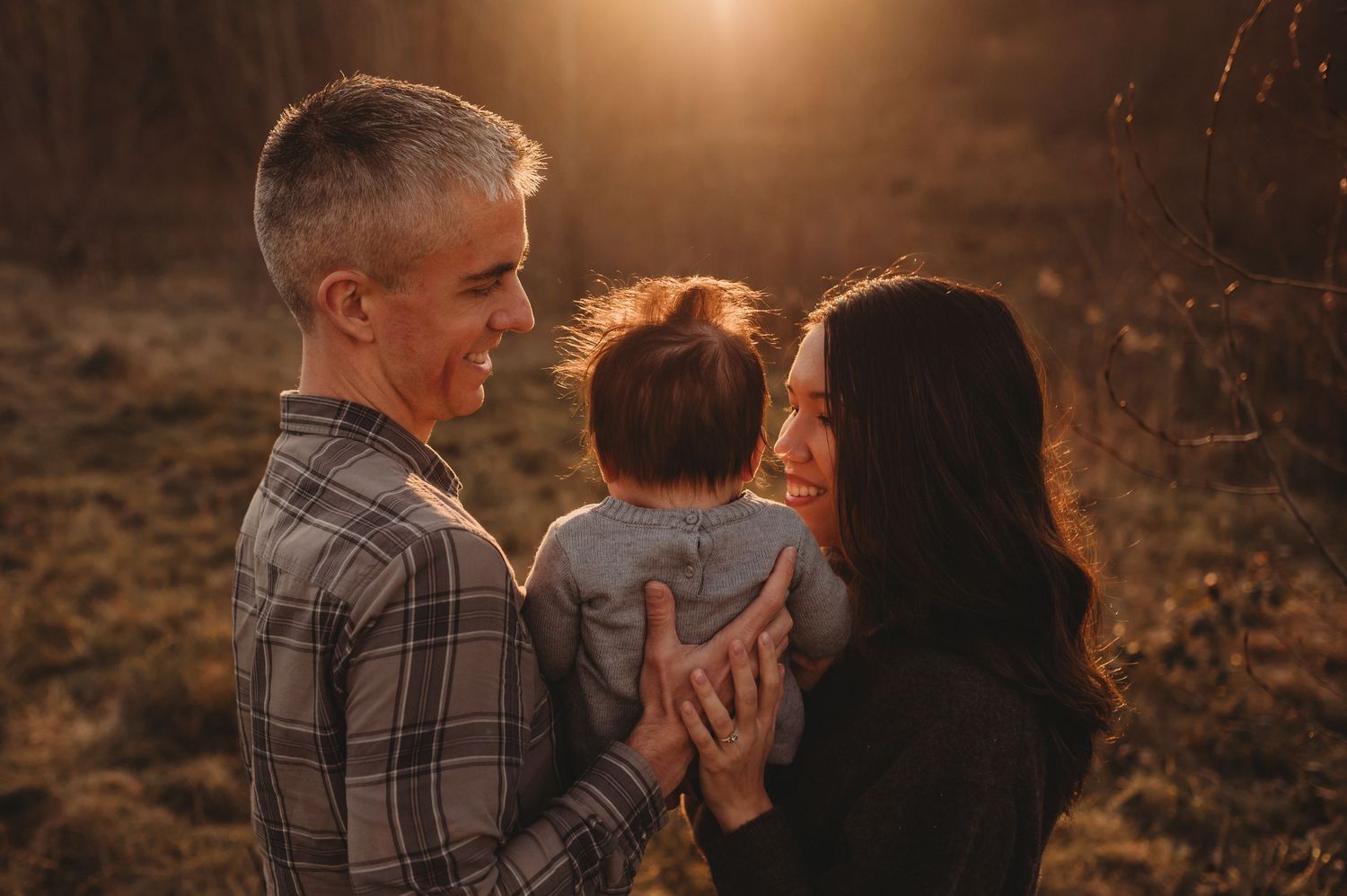 Smiling couple holding baby together in warm golden sunset light during outdoor family photo session.