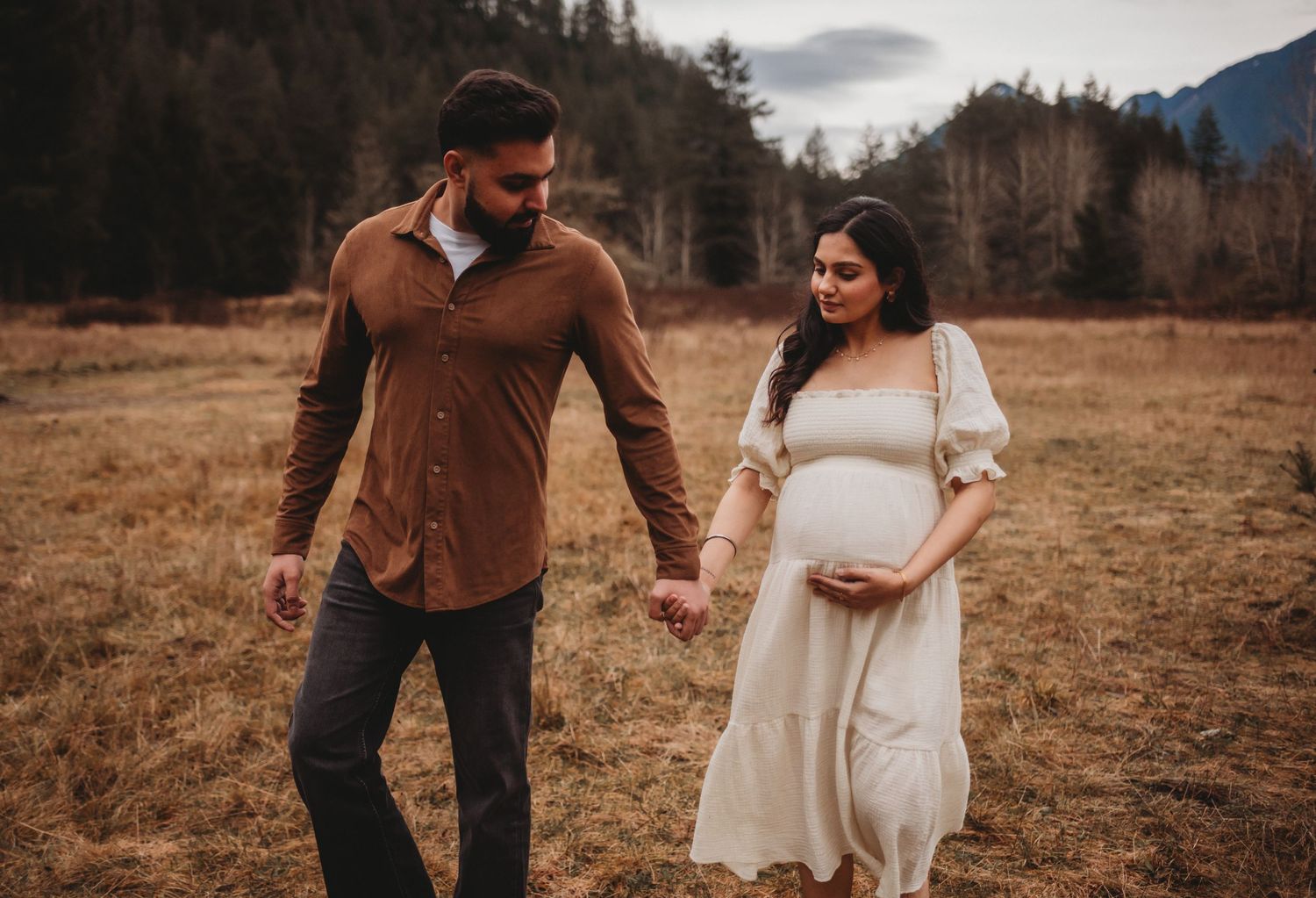 Man in brown shirt walking with pregnant partner in white dress through golden meadow.