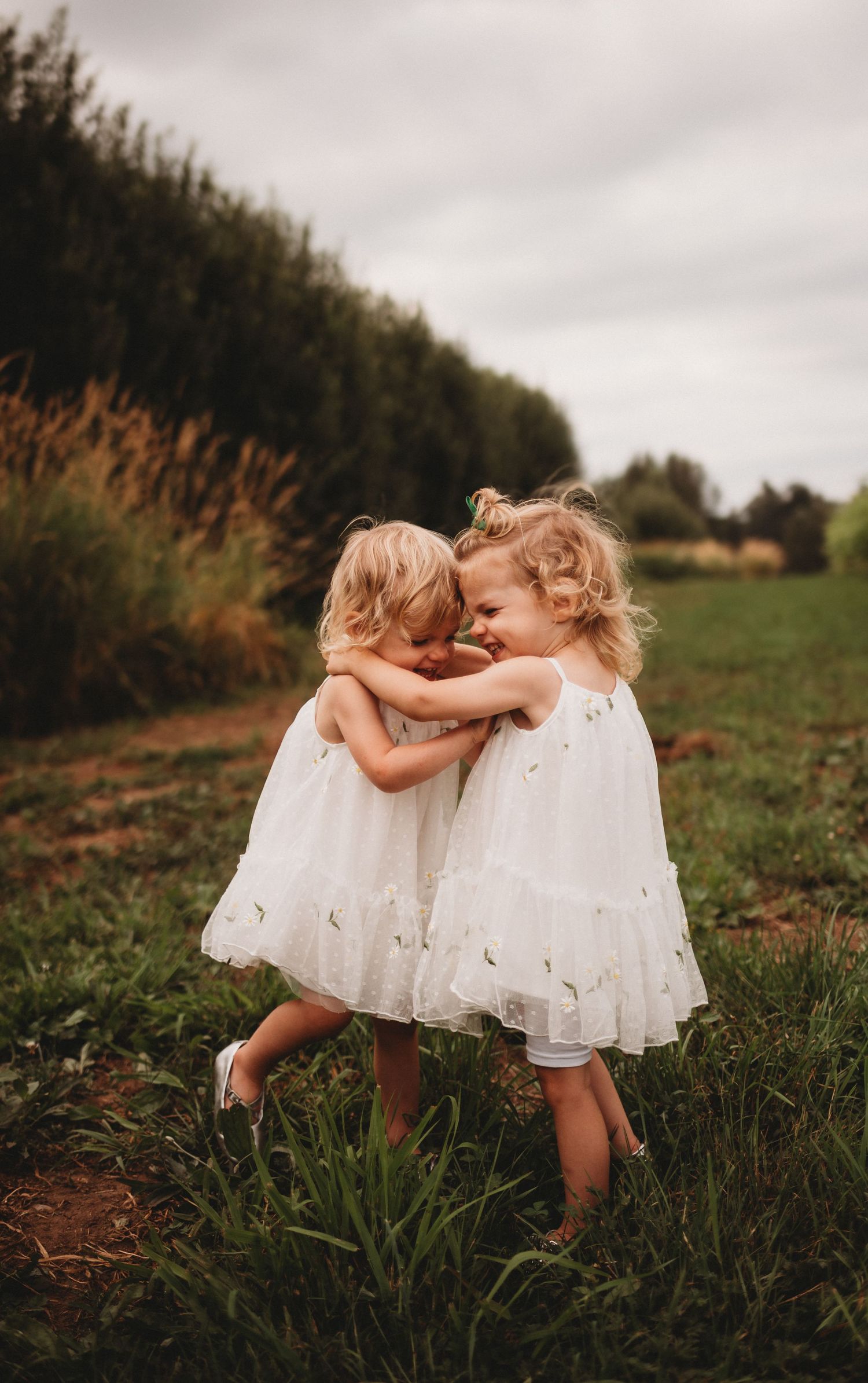 Two little girls in white dresses hug each other in a green field.