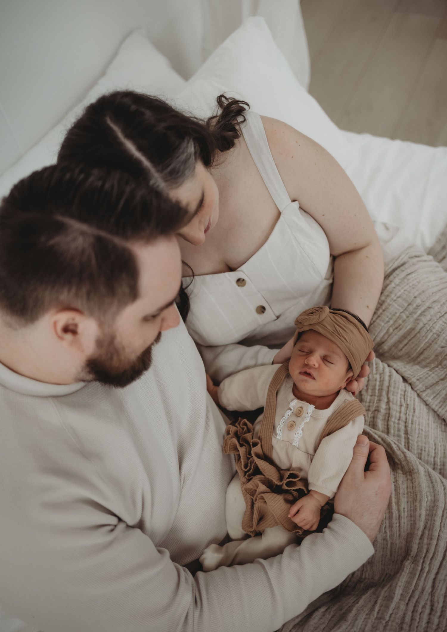 New parents lying together on a white bed, tenderly looking at their sleeping newborn baby.
