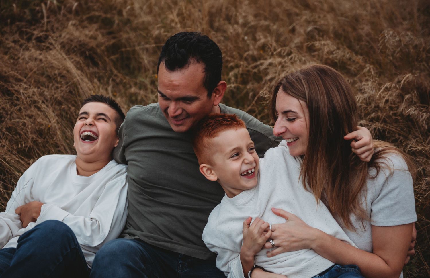 Happy family of four laughing together while sitting in golden autumn grass field.