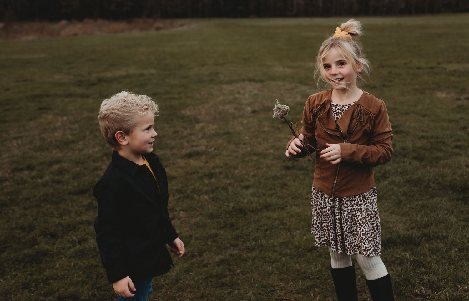 Two young children stand in a green field, with a girl in a brown fringe jacket holding a wildflower.