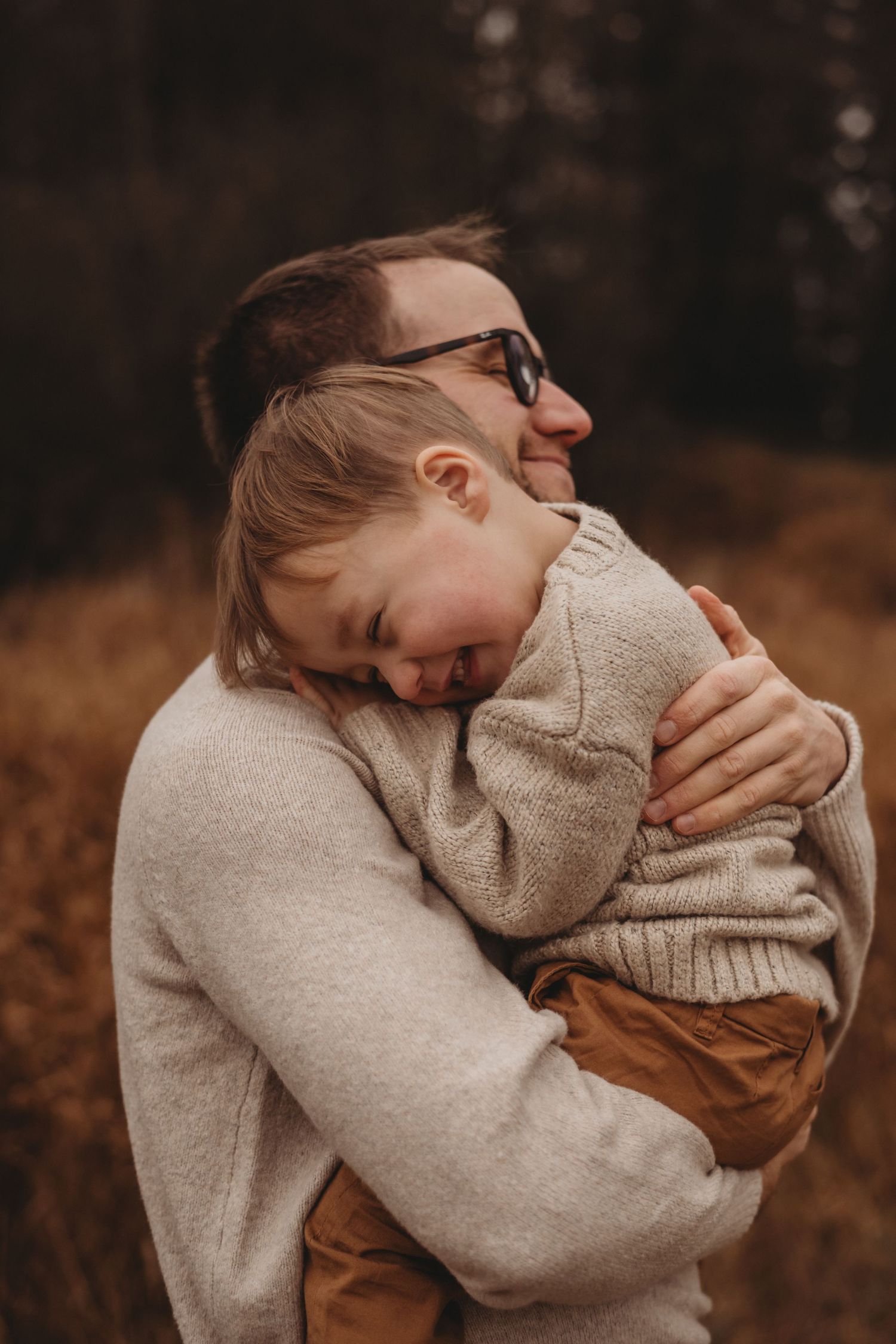 Father in cream sweater hugging young child tightly in outdoor fall setting.