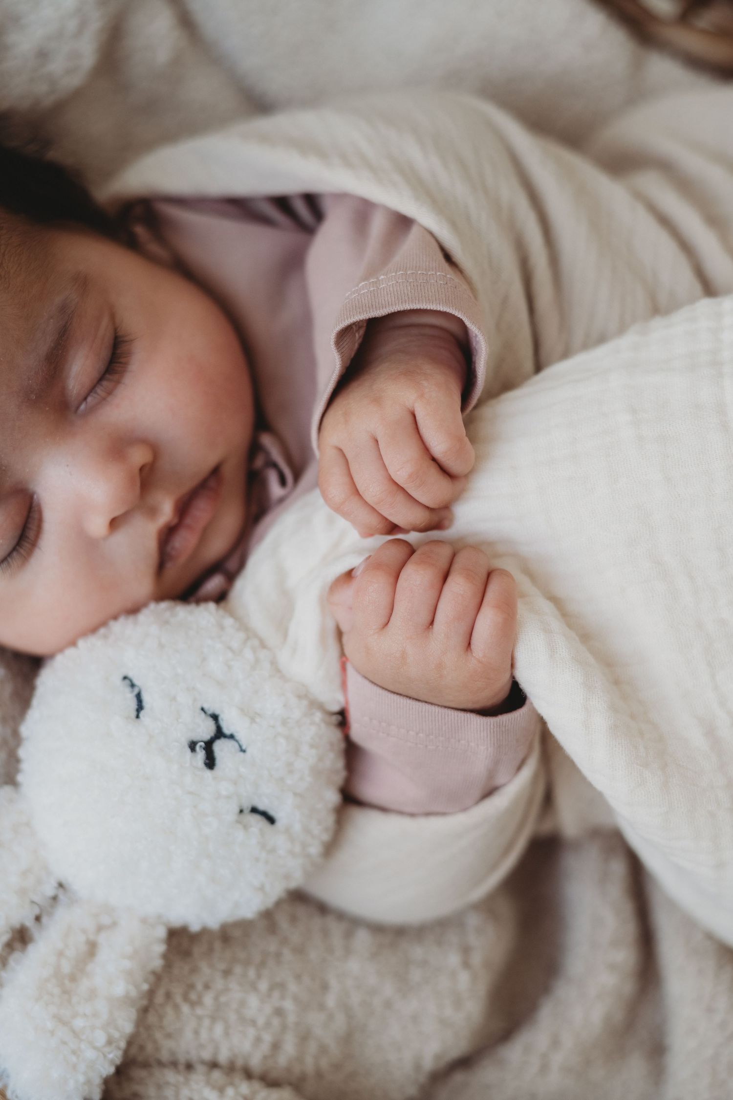 Sleeping newborn baby in pink outfit clutching a white stuffed bunny toy wrapped in a soft blanket.