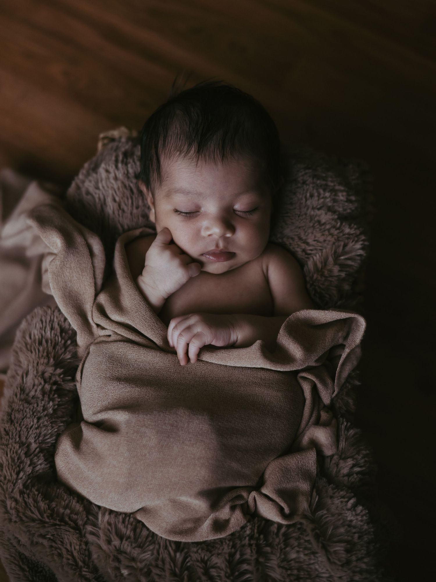 Newborn baby sleeping peacefully in brown wrap nestled in fluffy basket on wood floor.