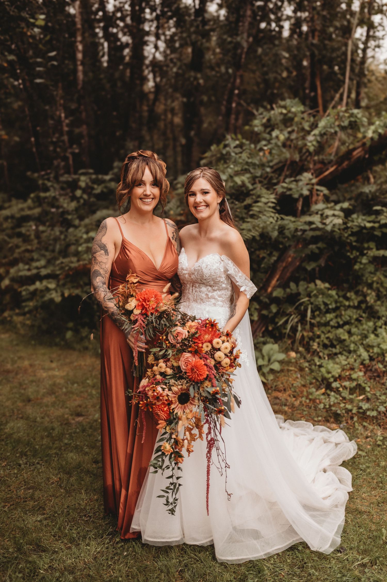 Bride in lace gown poses with bridesmaid in rust orange dress holding cascading fall floral bouquet.
