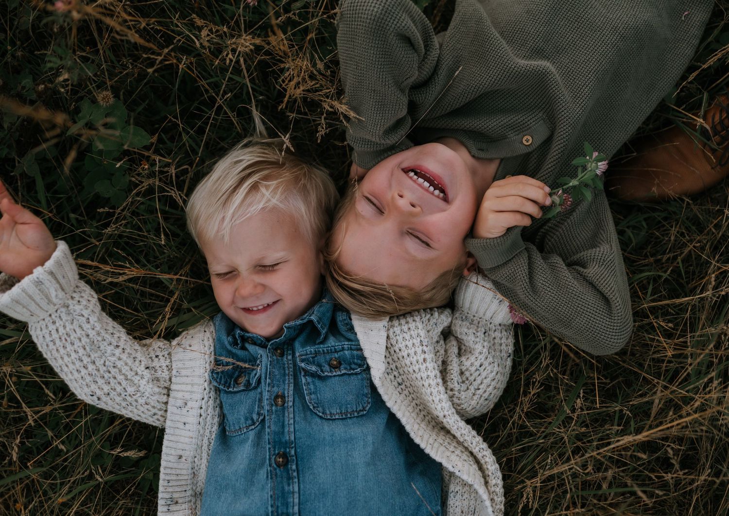 Two young siblings laugh and lay together on grass, sharing a joyful candid moment during a family session.