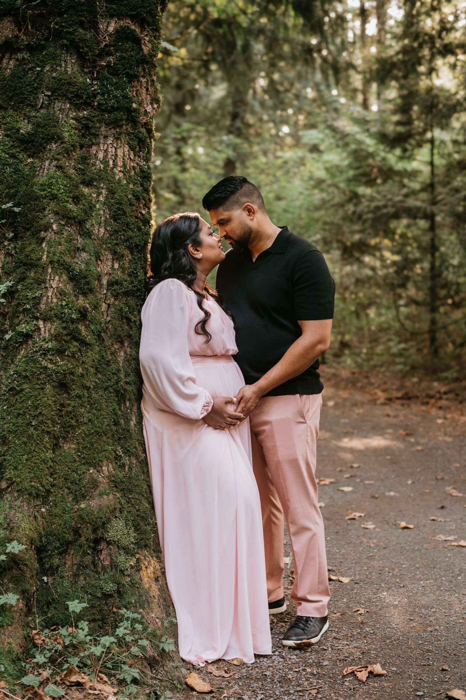 Pregnant woman in pink dress and partner sharing romantic moment beside mossy tree in forest.