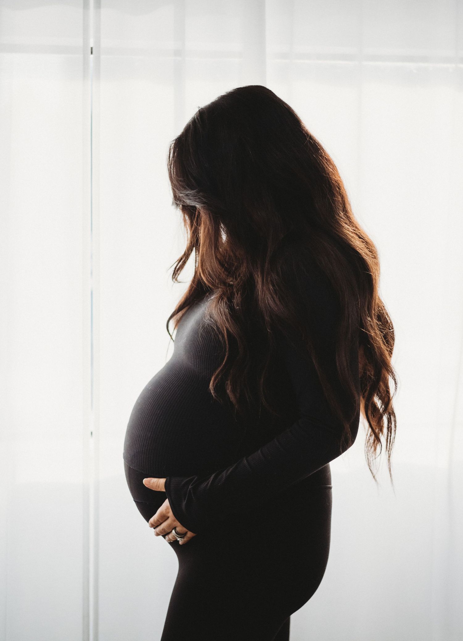 Pregnant woman in black dress holding baby bump in silhouette by window.