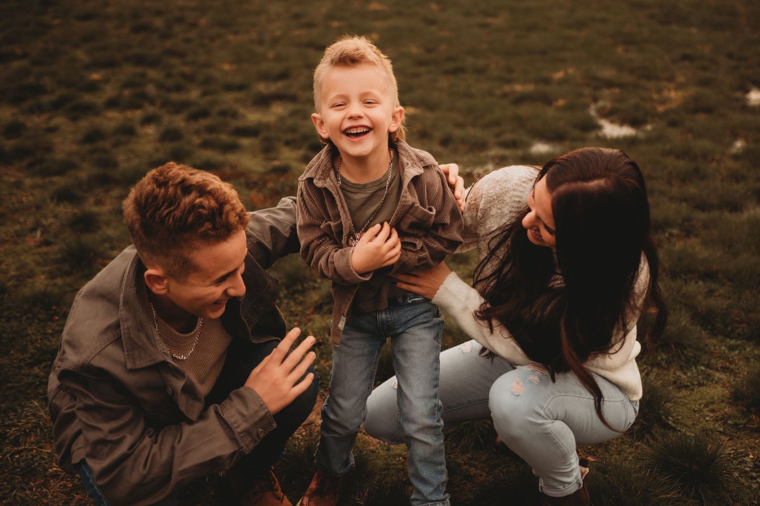 Parents kneeling beside their joyful laughing boy on a green outdoor field.