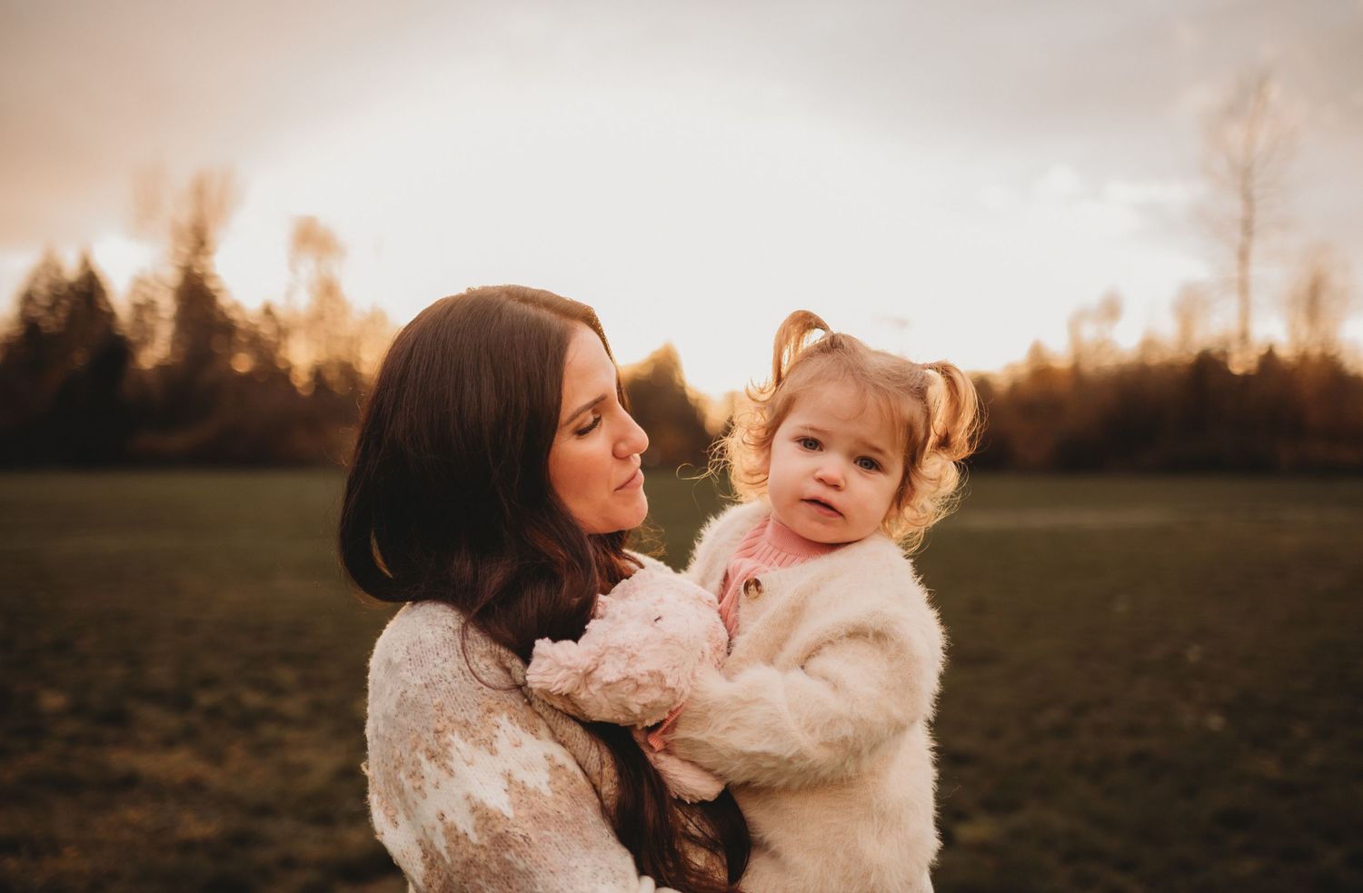 A dark-haired mother holds her toddler daughter in a sunlit field at golden hour.