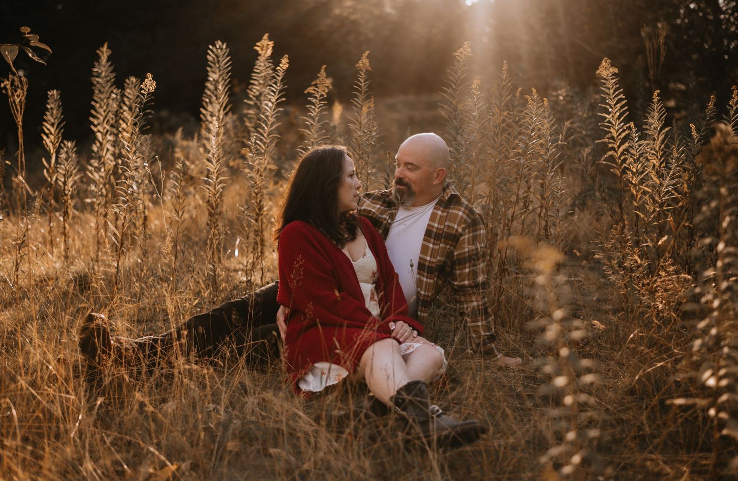 Couple sitting together in golden field at sunset, holding hands among tall grasses.