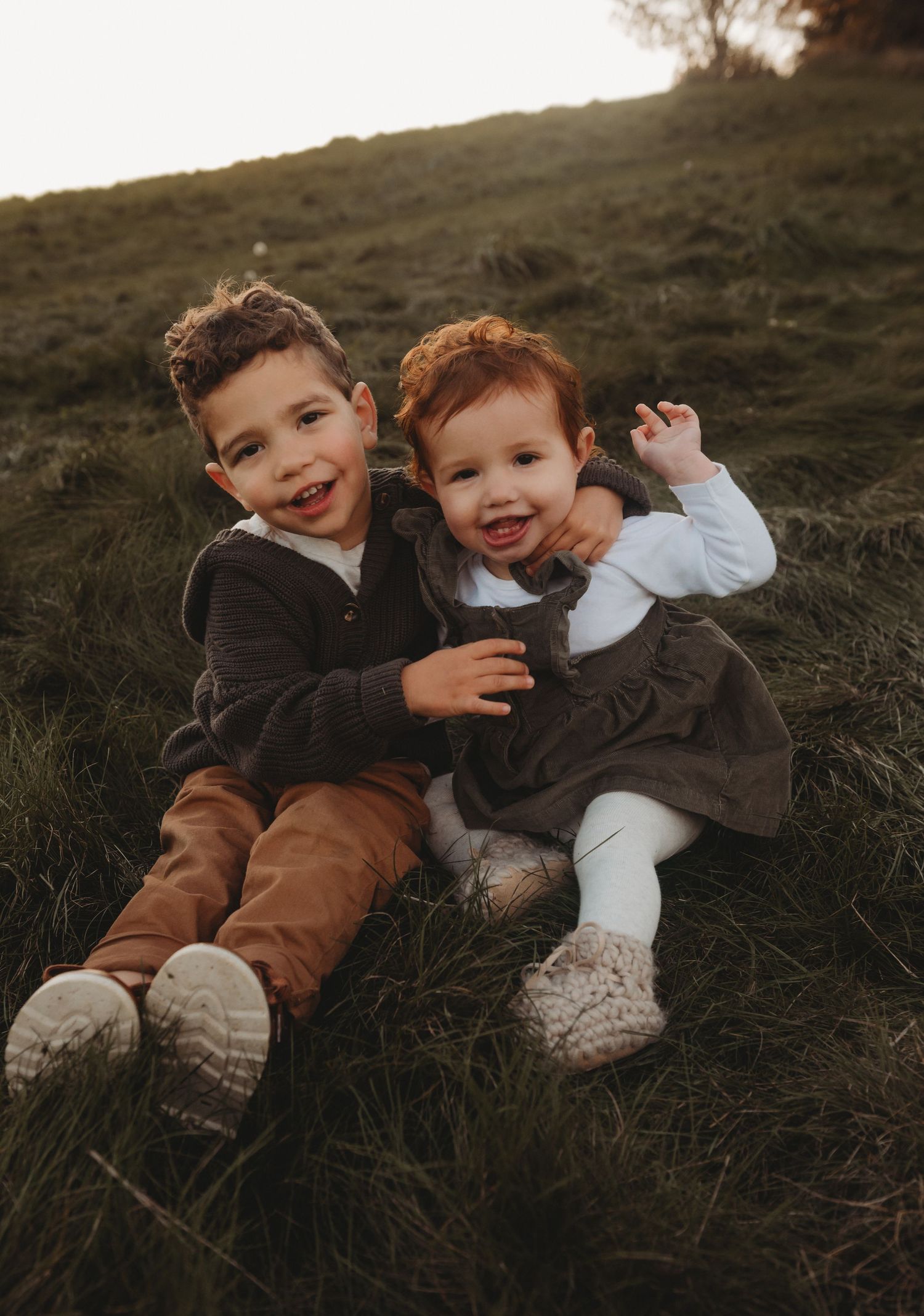 Two adorable young siblings smile and pose together while sitting on a grassy hillside in autumn light.