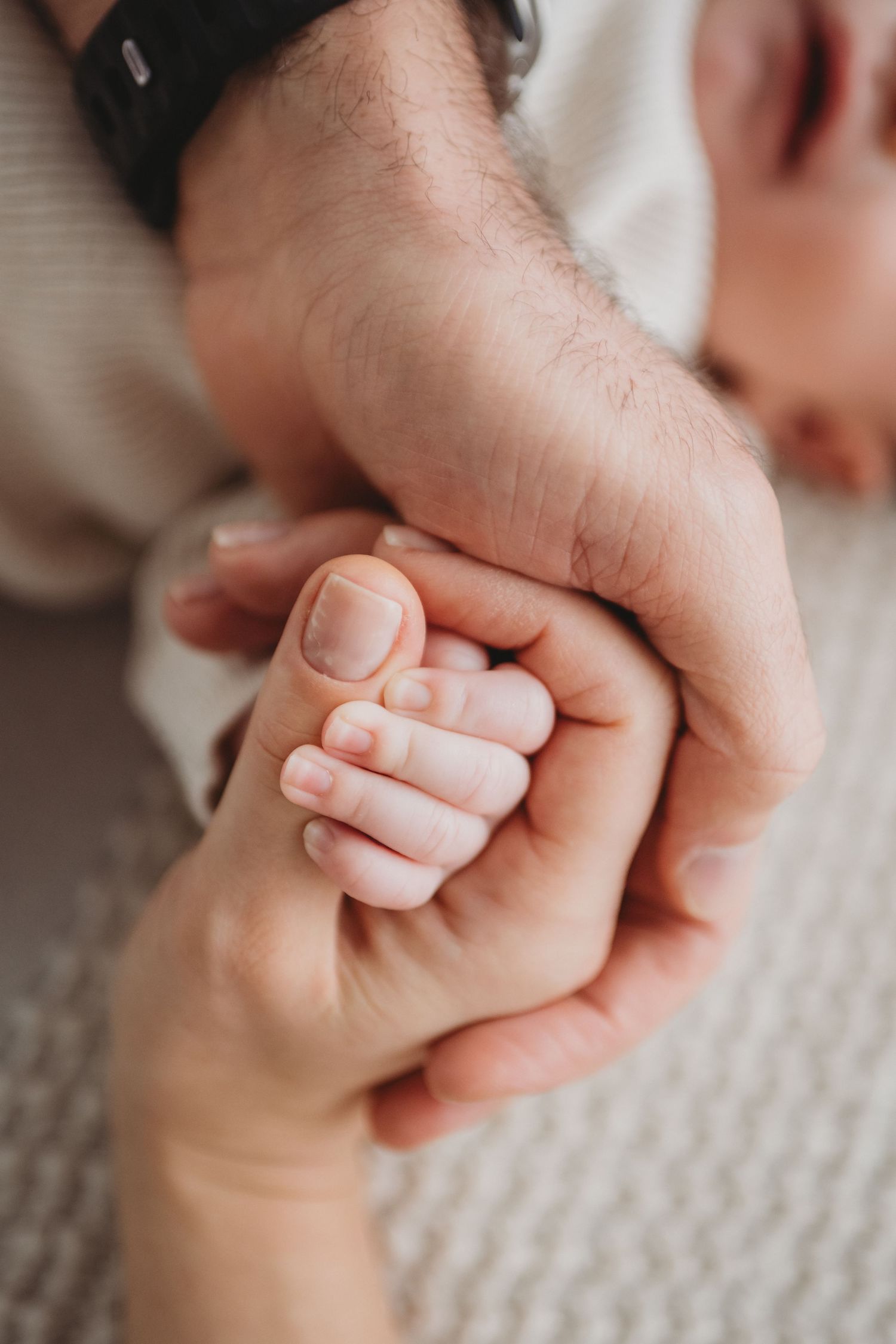 Close-up of a newborn baby gripping parents fingers with tiny hand on a white blanket.