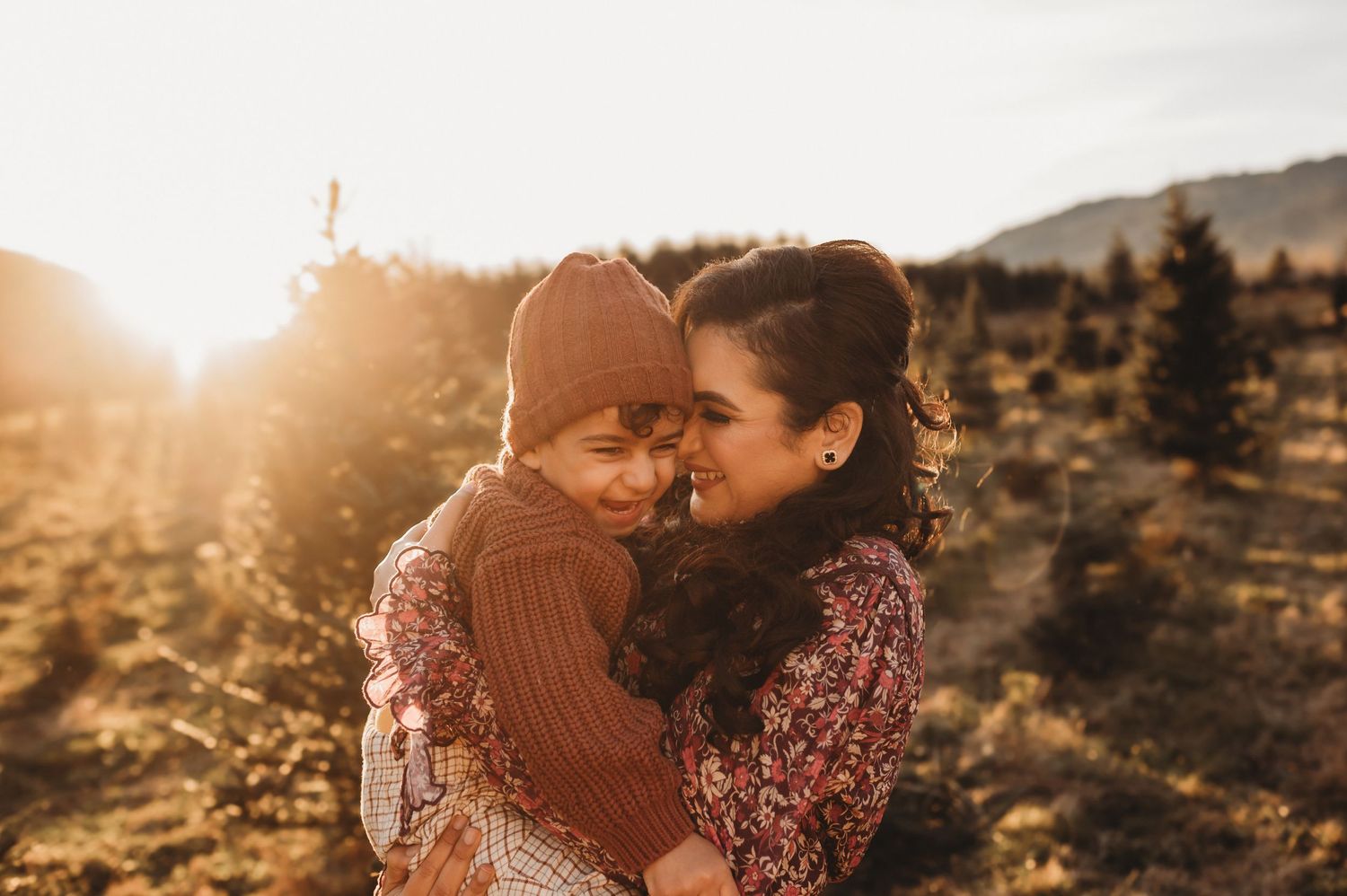 Mother hugging young child in warm sunset light at Christmas tree farm.
