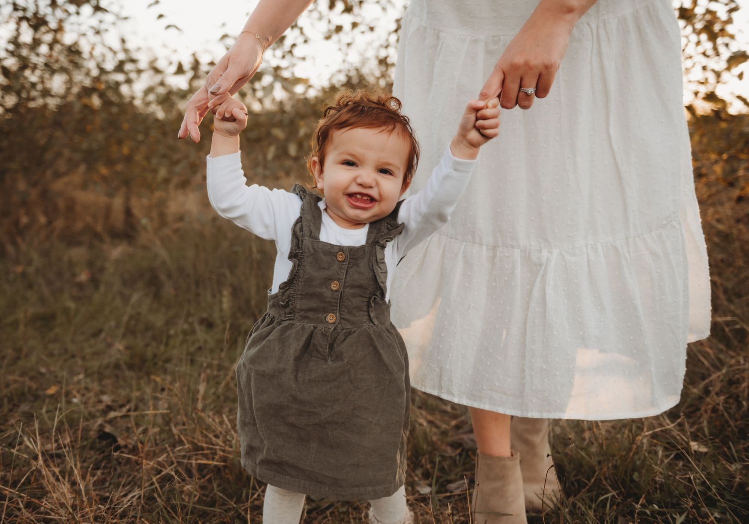 Baby girl smiling while mother holds her hands in field.