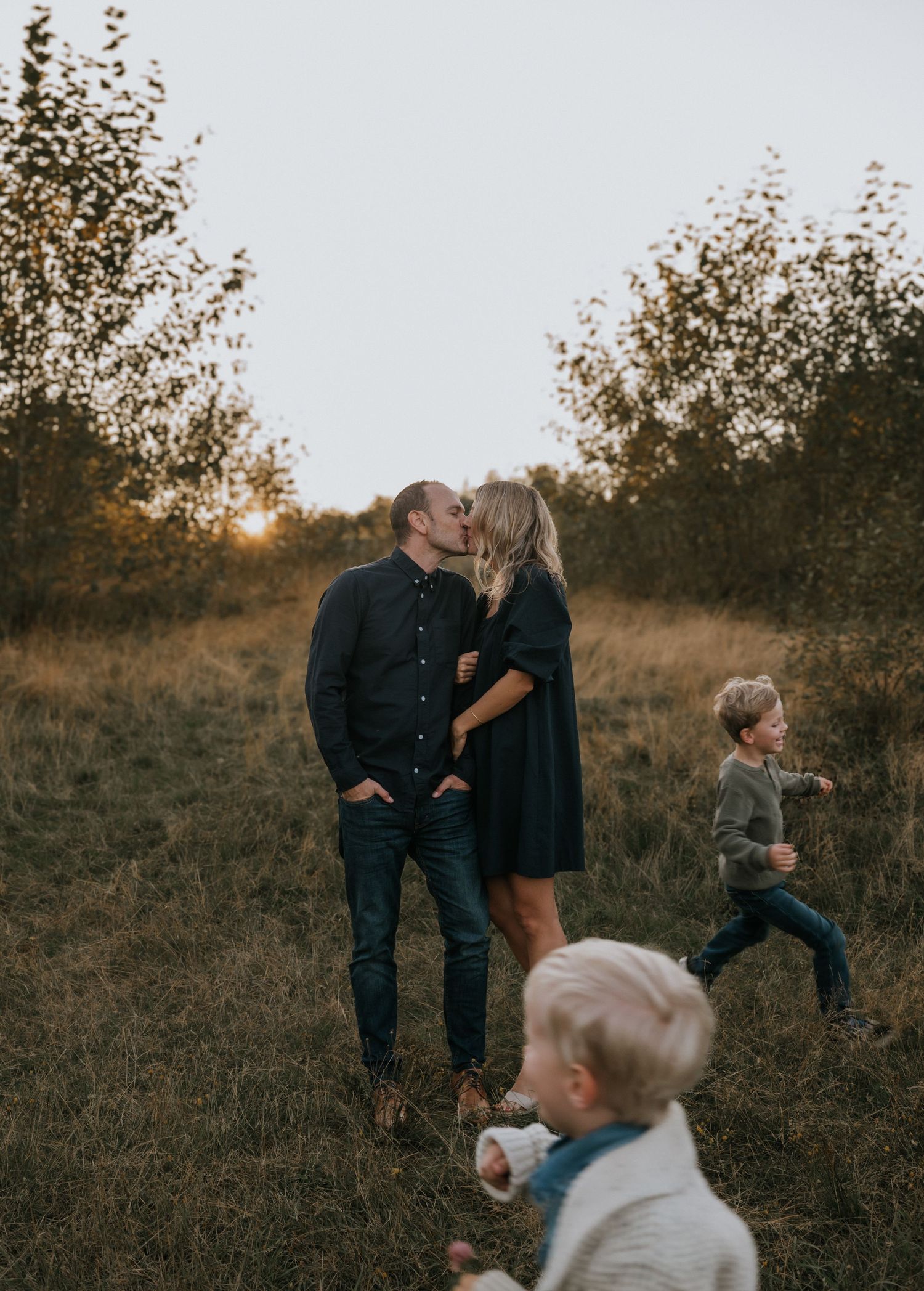 Couple sharing a kiss in a golden field at sunset while two young boys play around them.