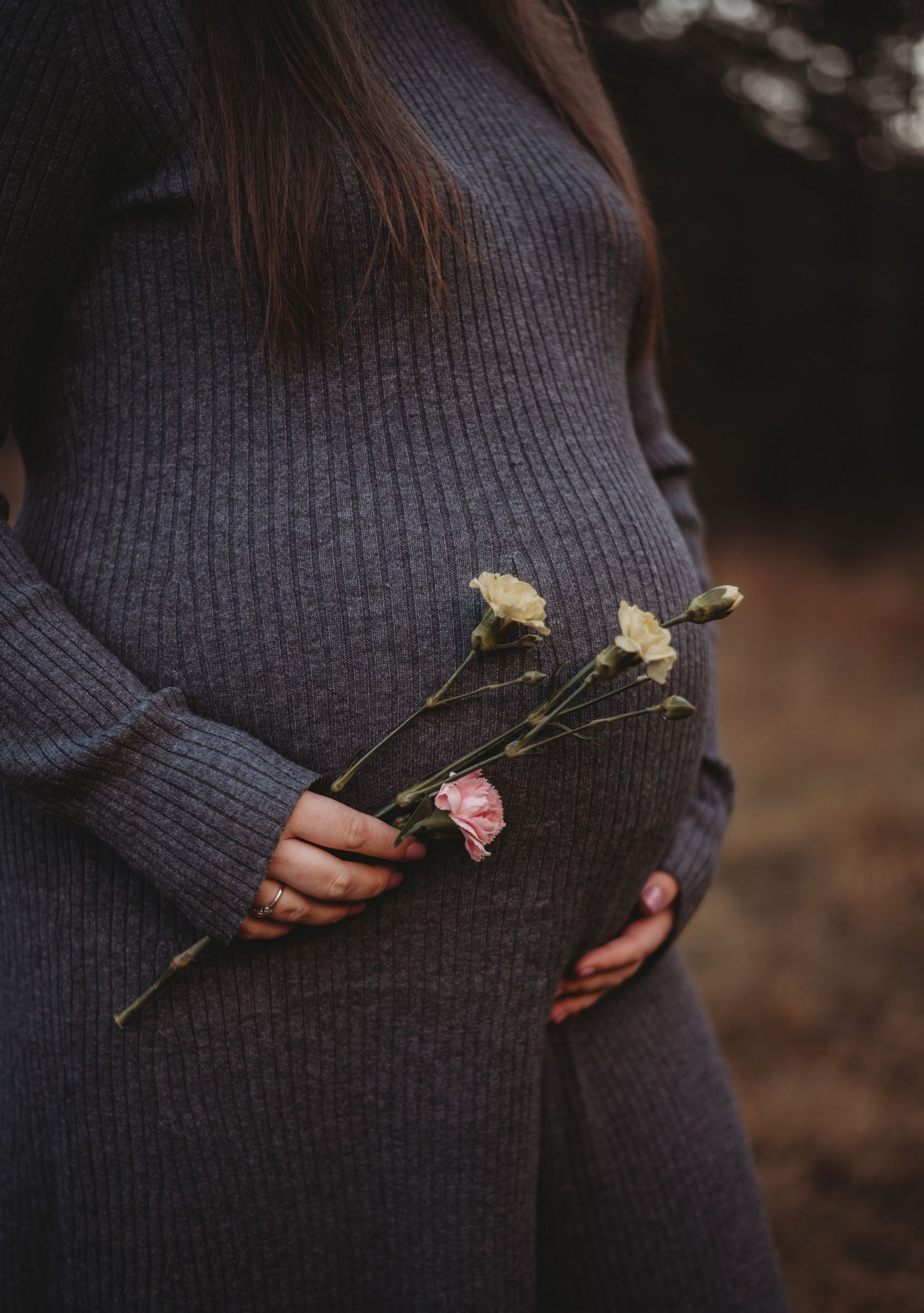 Close-up of pregnant woman in gray knit dress holding delicate pink and white flowers.