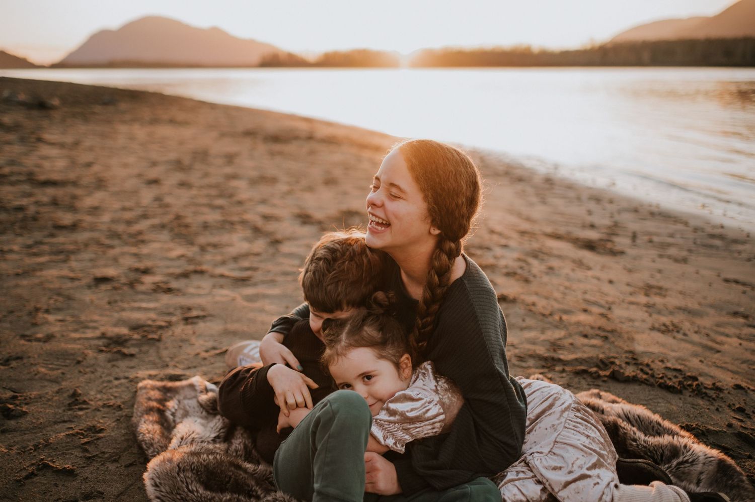 Joyful mother laughing while hugging two children on sandy beach at golden sunset by the water.