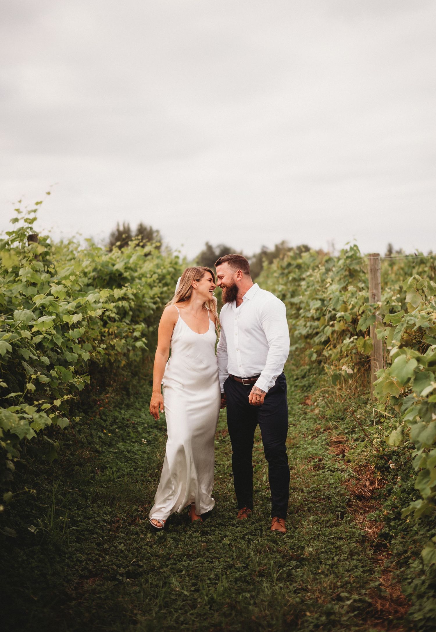 A couple in wedding attire shares a romantic moment walking through a lush green vineyard under cloudy skies.