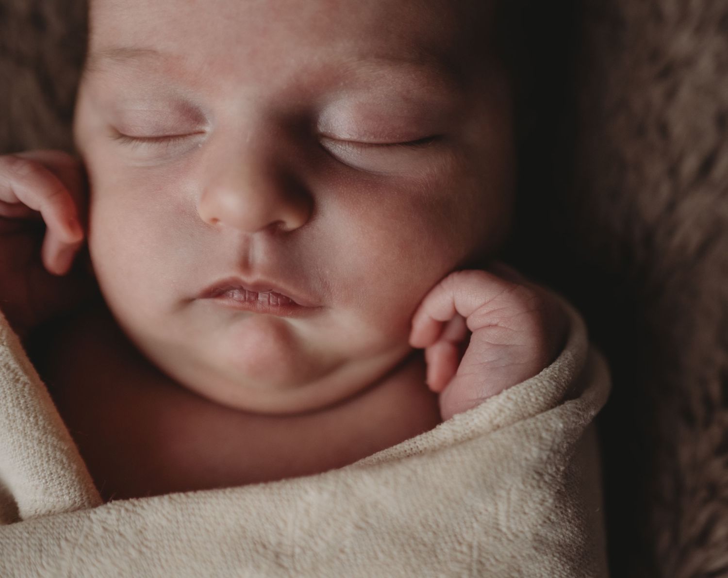 A close-up of a peacefully sleeping newborn baby swaddled in a soft cream blanket.