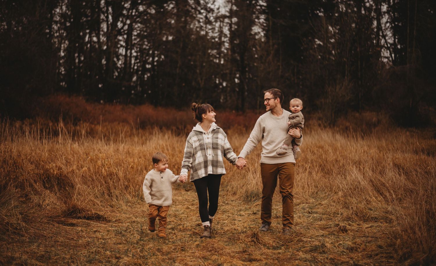 Happy family of four walking hand in hand through golden autumn field near dark forest.