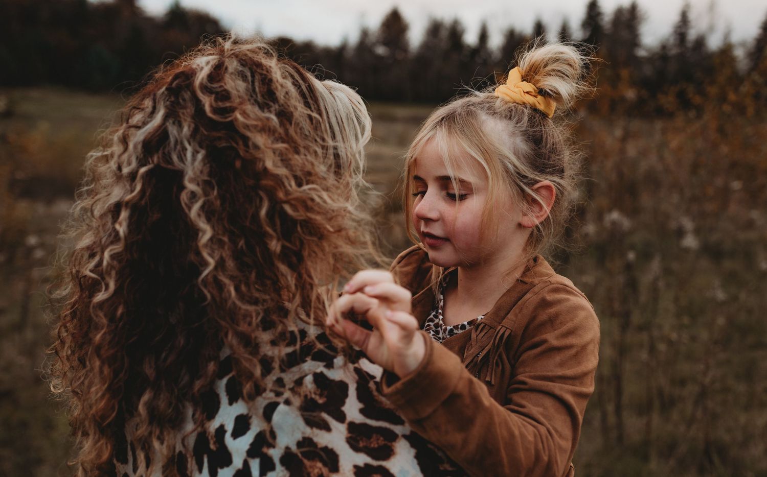 Young blonde girl in brown jacket holding hands with curly-haired woman in leopard print outdoors.