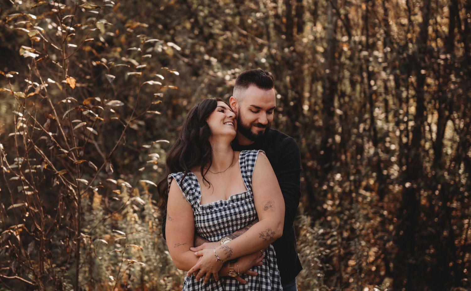 A couple laughs together in a golden autumn forest, the man nuzzling the smiling woman in a gingham dress.