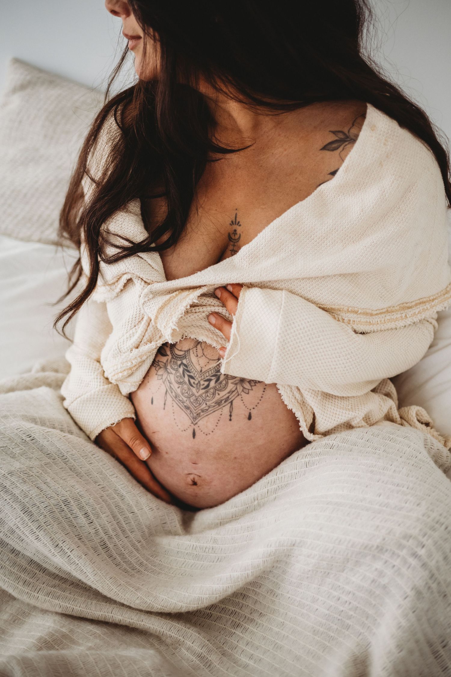 Close-up of pregnant belly with mandala tattoo wrapped in cream blanket.
