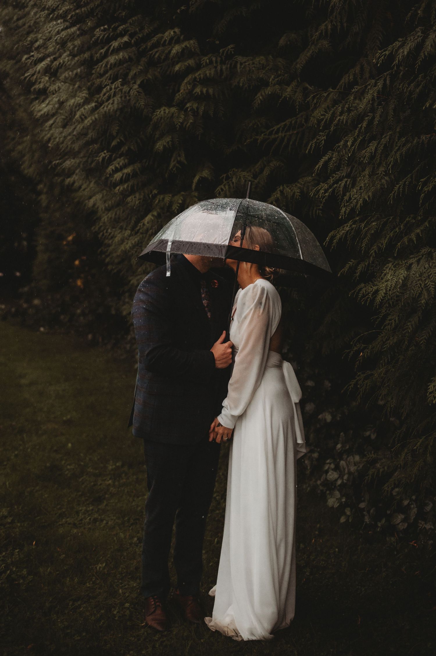 Newlyweds embracing under a clear umbrella in a dark lush garden on their rainy wedding day.