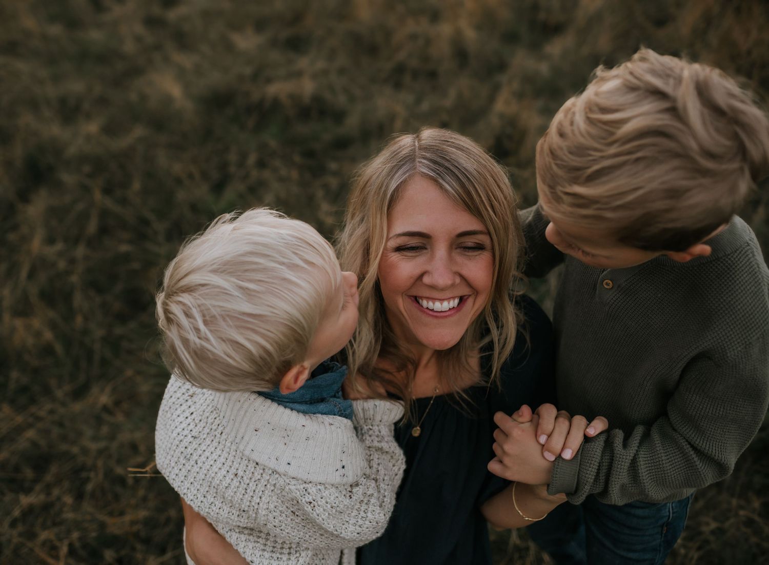 Smiling mother embraced by two young blonde children in a field.