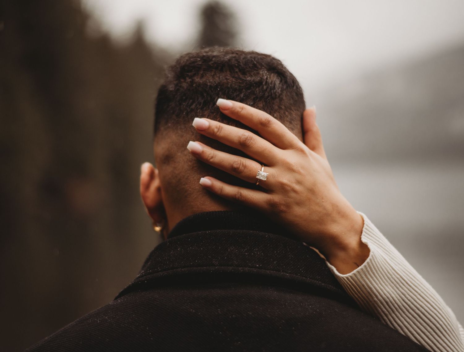 Woman with engagement ring gently placing hand on back of mans head outdoors.