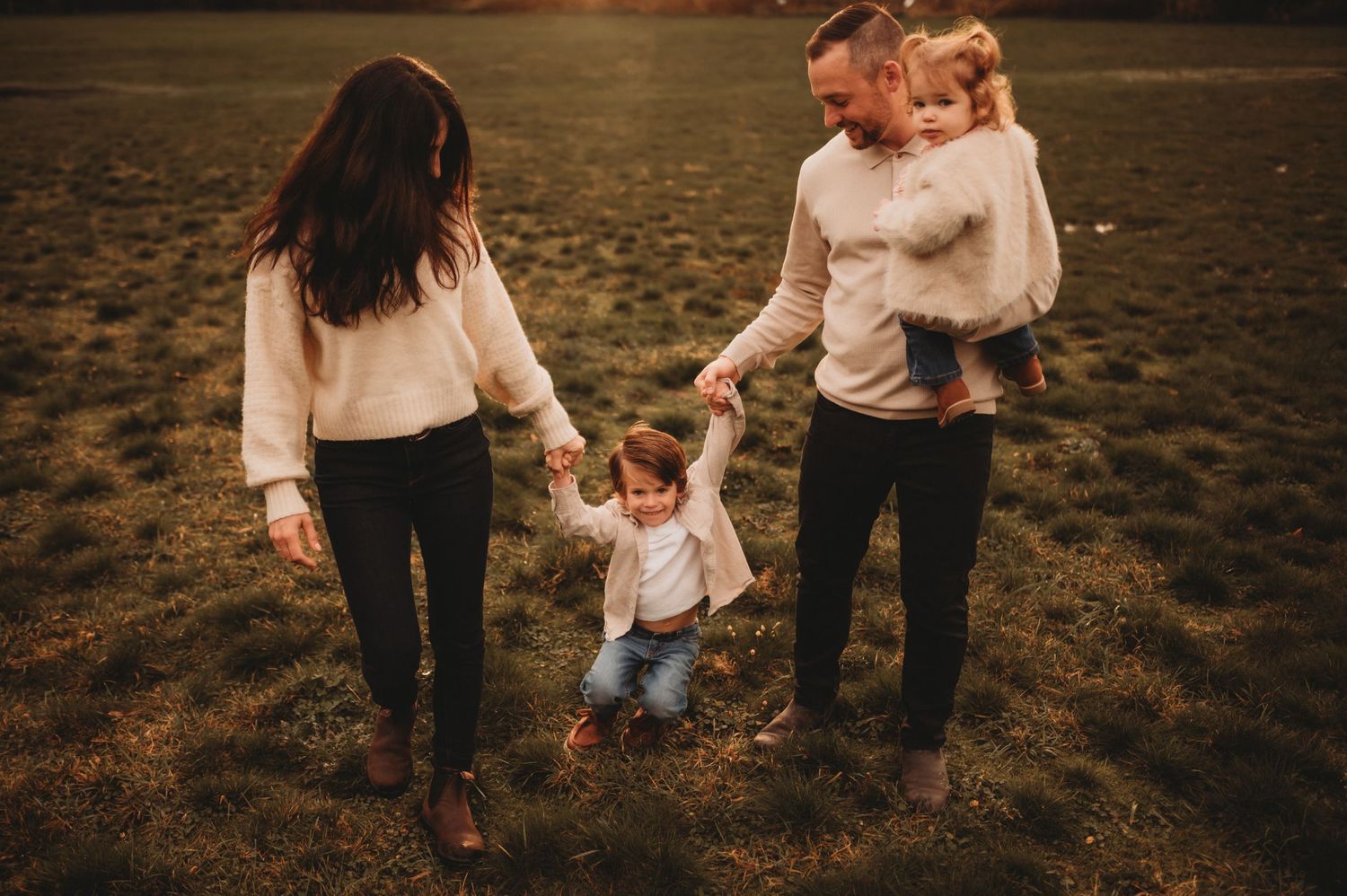 Family of four holding hands and walking together across a grassy field at sunset.